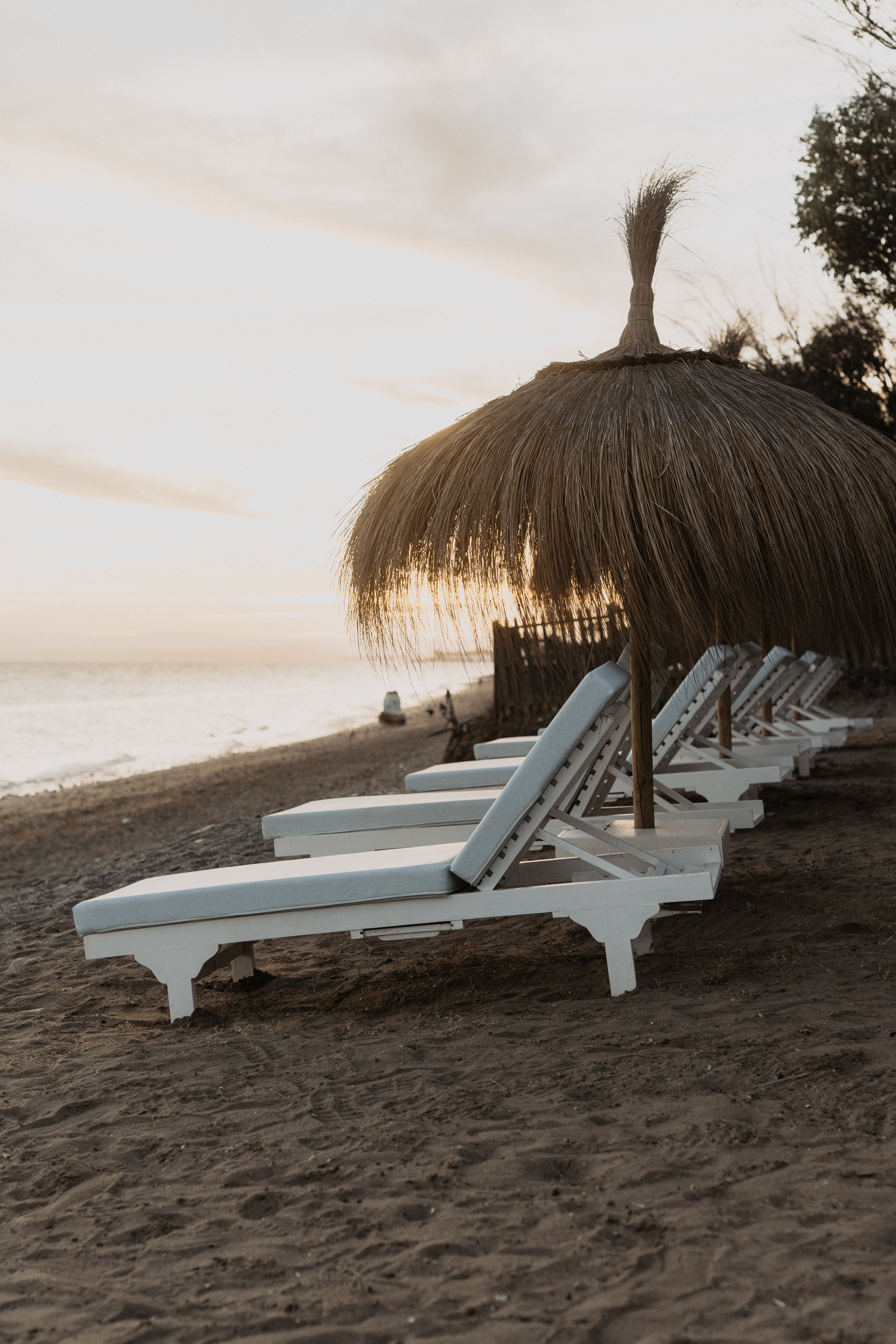 Beach with white lounge chairs under a thatched umbrella at sunset or sunrise.