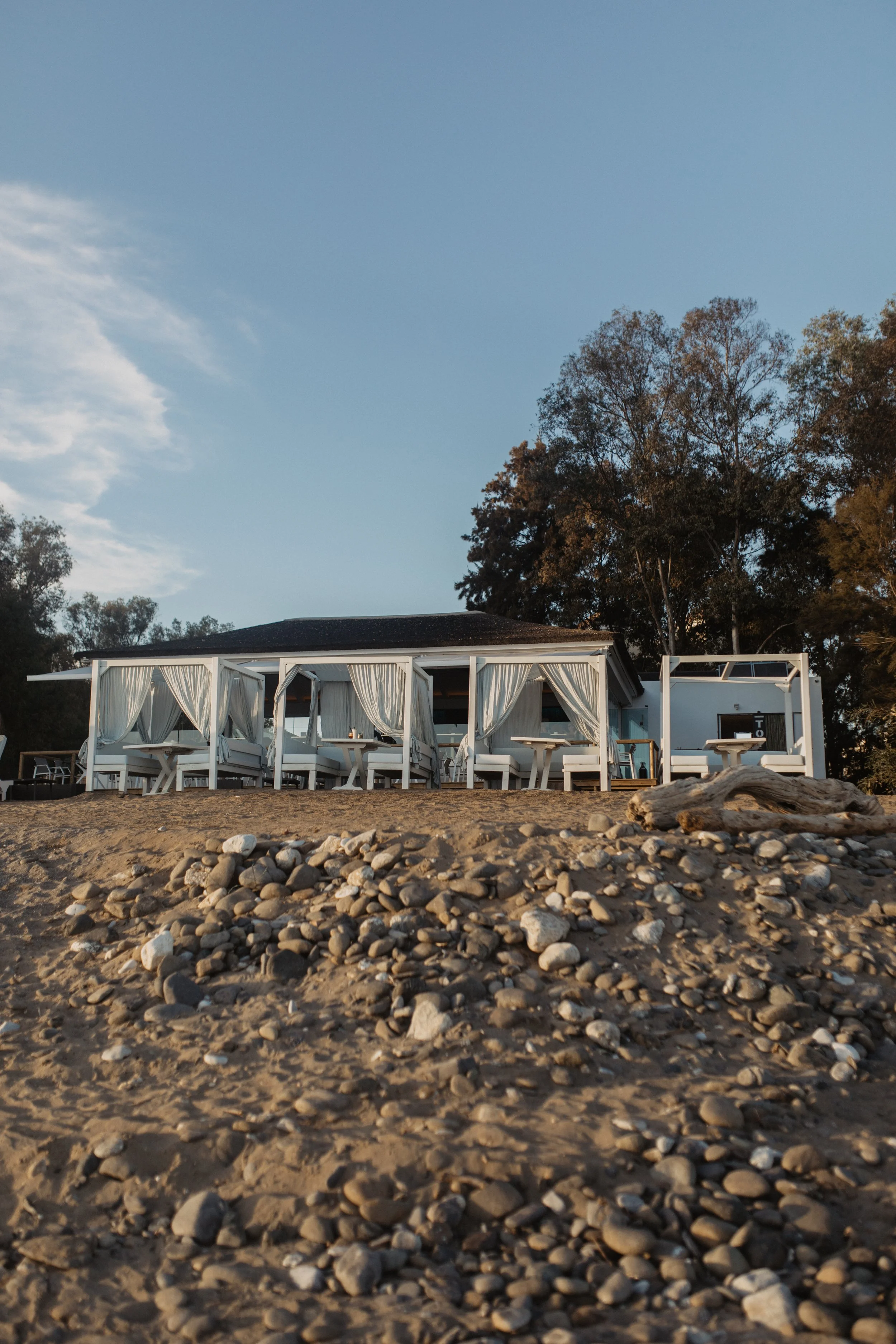 Beachside cabana with white curtains and seating, on a sandy and rocky shore with trees in the background under a clear blue sky.