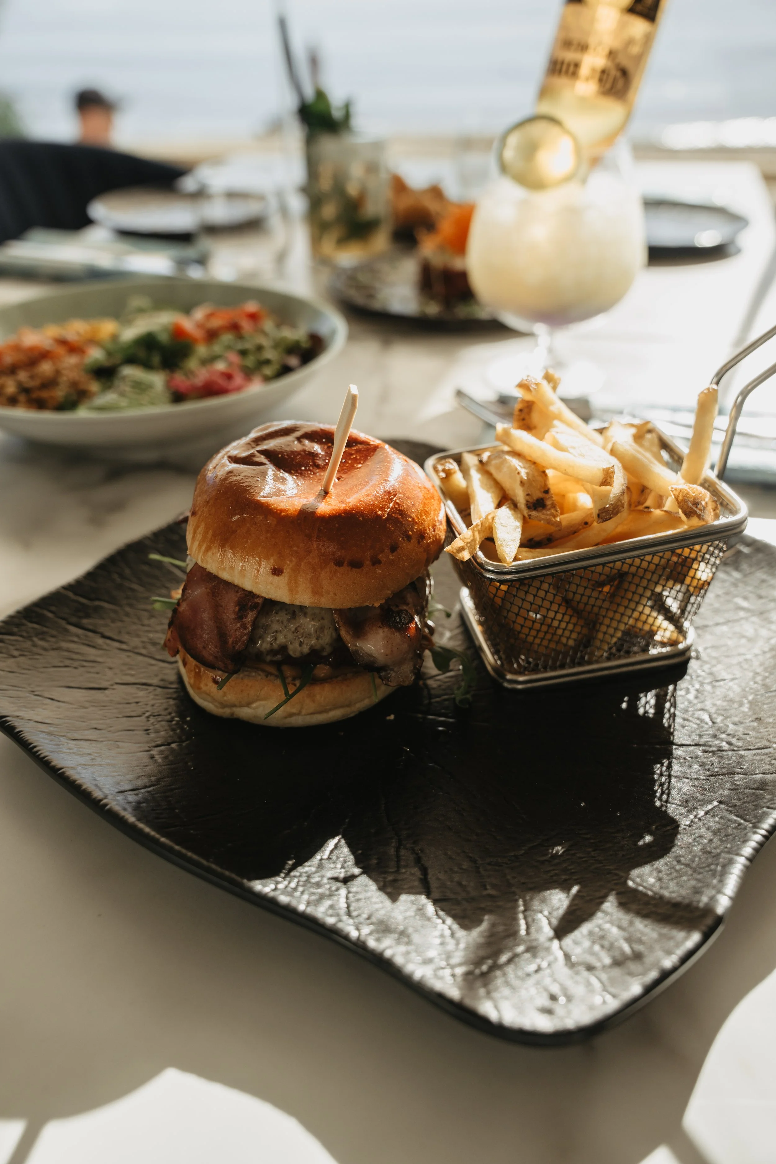 Close-up of a burger with bacon and greens served with a side of French fries on a black plate, with a salad and a drink in the background.