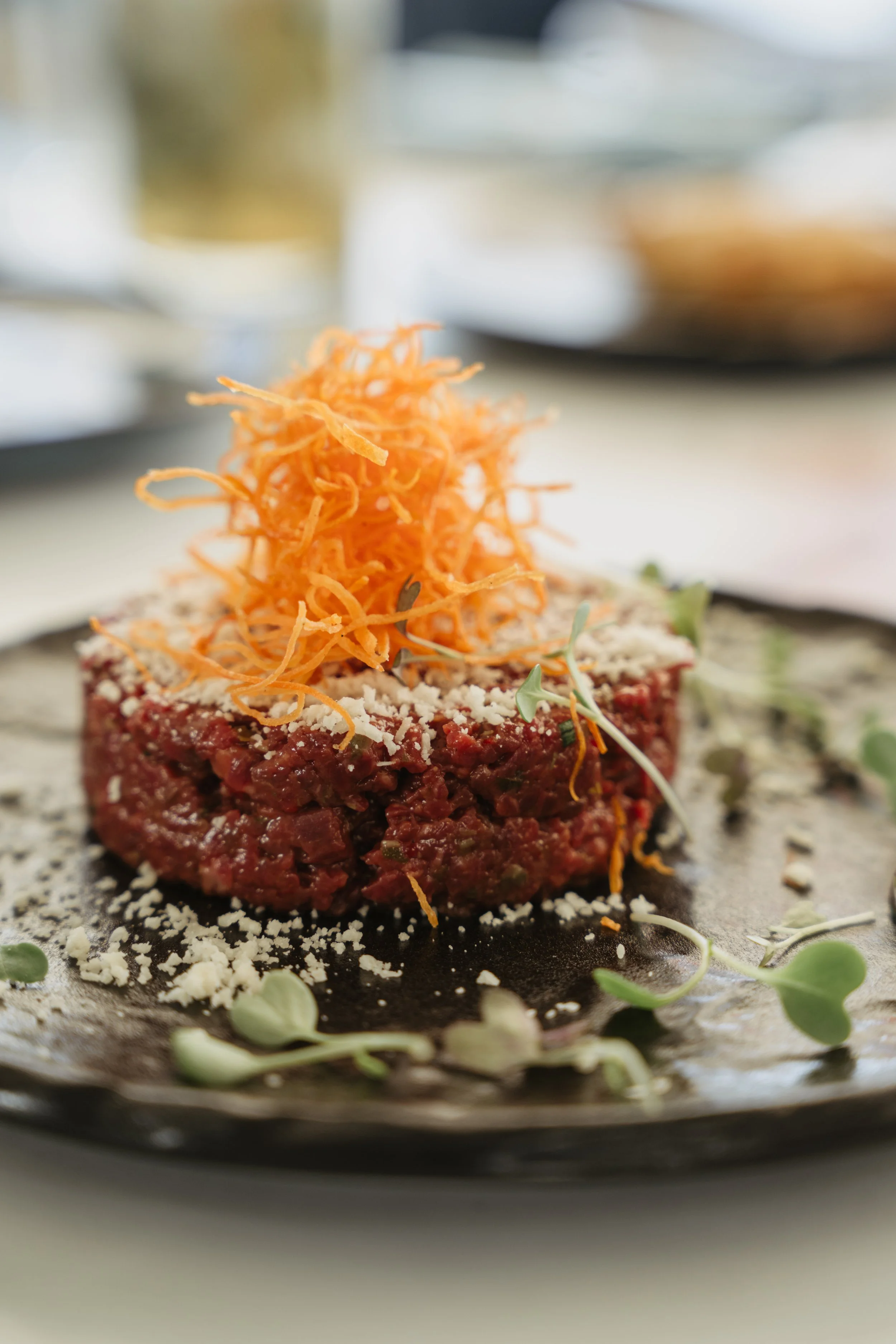A dish of seasoned raw beef tartare topped with shredded carrots and garnished with microgreens, served on a black plate with a sprinkle of grated cheese.