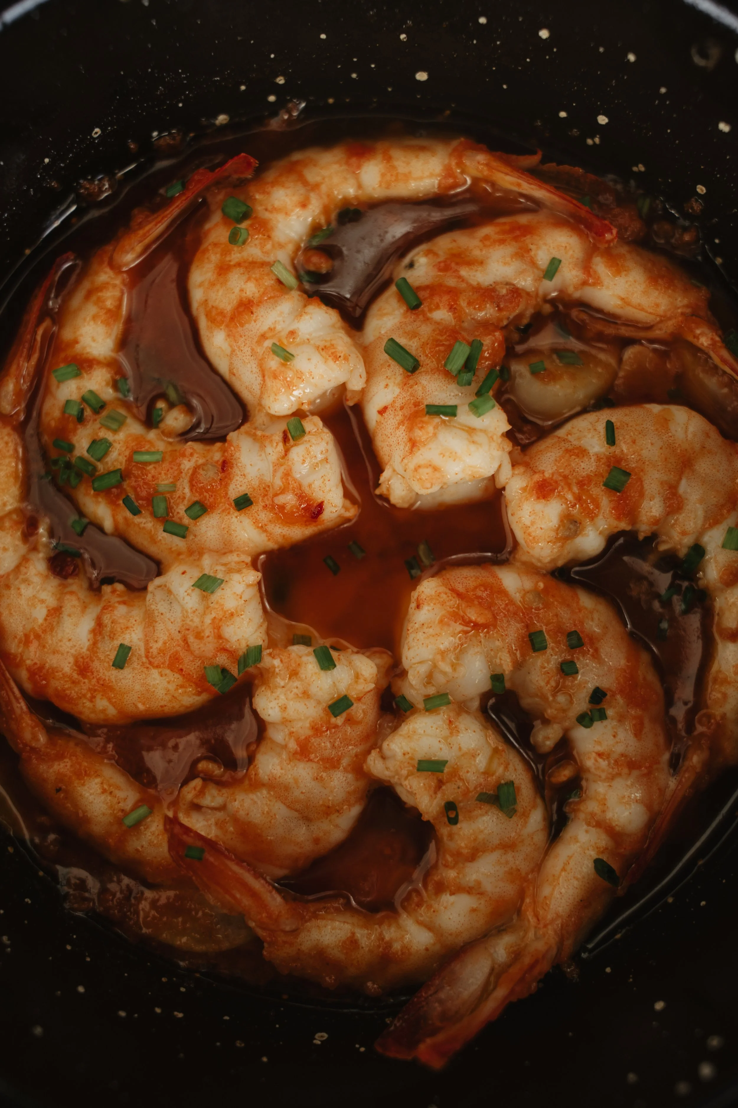 Cooked shrimp with sauce and chopped chives in a black bowl.