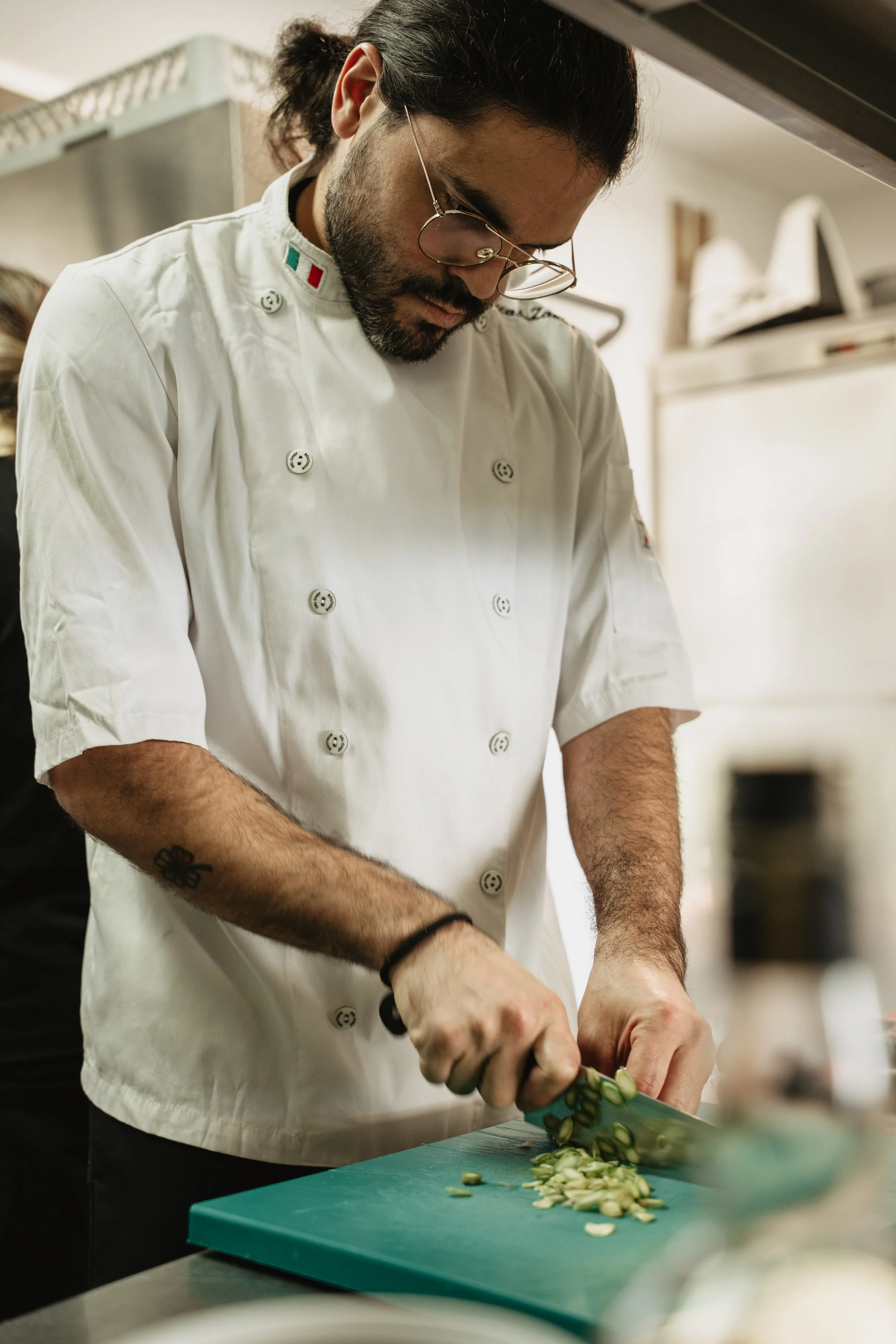 A chef in a white uniform and glasses chopping green vegetables on a green cutting board in a kitchen.
