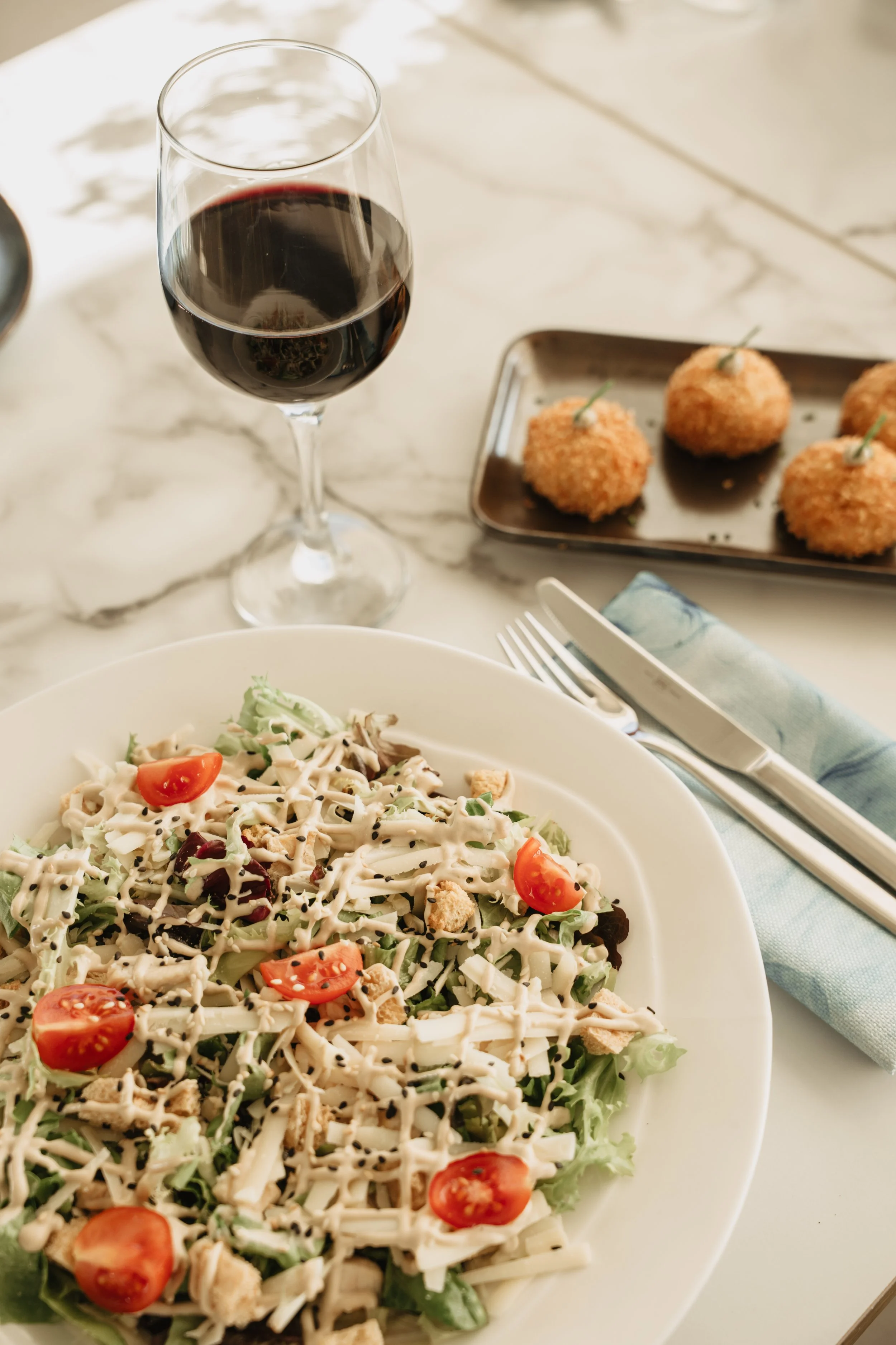 A plate of salad with cherry tomatoes, shredded cheese, and dressing, a glass of red wine, and a tray with three fried appetizers on a marble table.