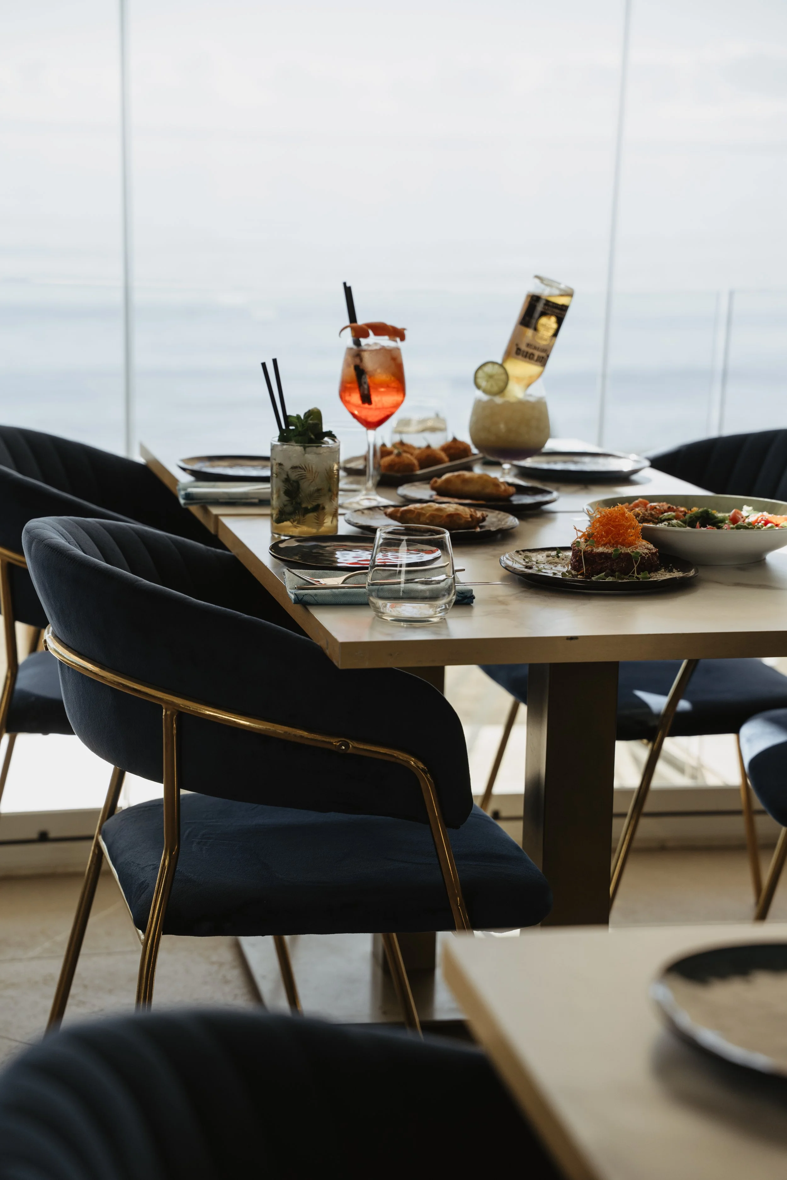 A dining table set with various dishes and colorful cocktails with a view of water through large windows.