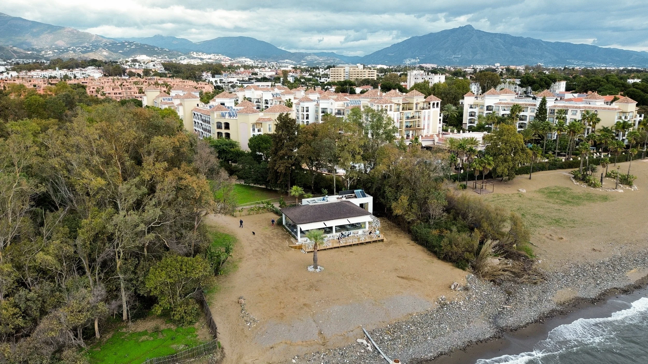 Aerial view of a beachside property with a house and patio, surrounded by trees, with residential buildings and mountains in the background.