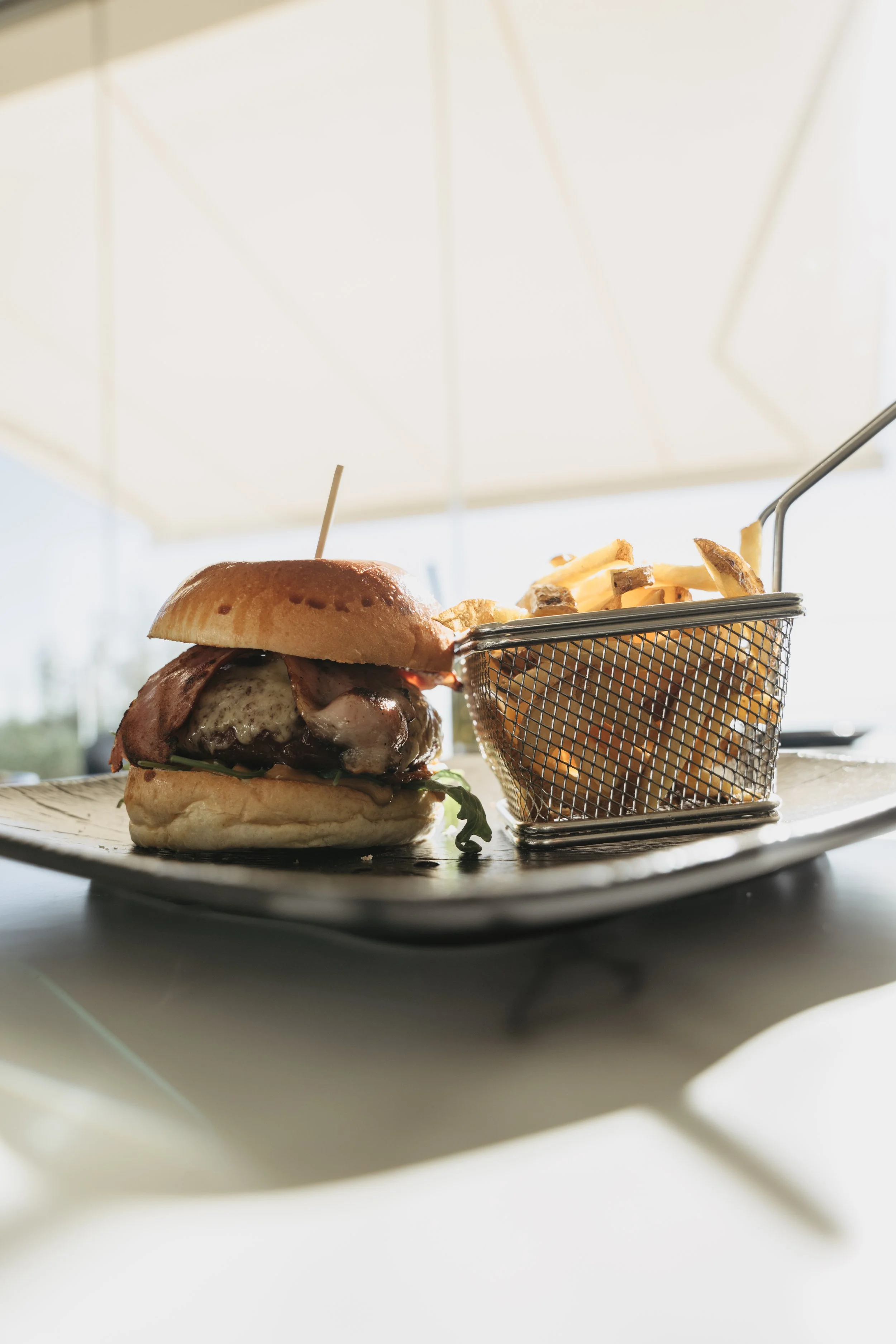 Close-up of a cheeseburger with bacon, lettuce, tomato, on a soft bun, served with a side of French fries on a metal basket, on a black serving tray under a white umbrella.