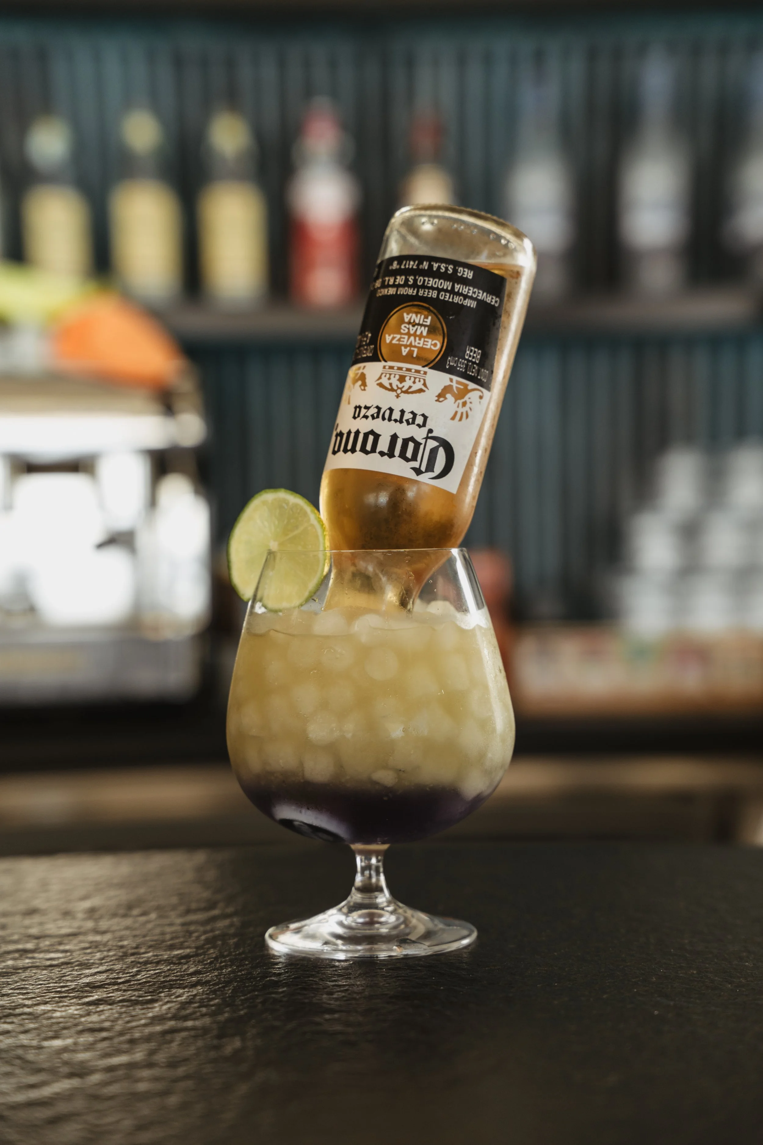 A cocktail glass filled with crushed ice, lime wedge, and a bottle of Corona beer inserted upside down, set on a bar counter with a blurred bar background.