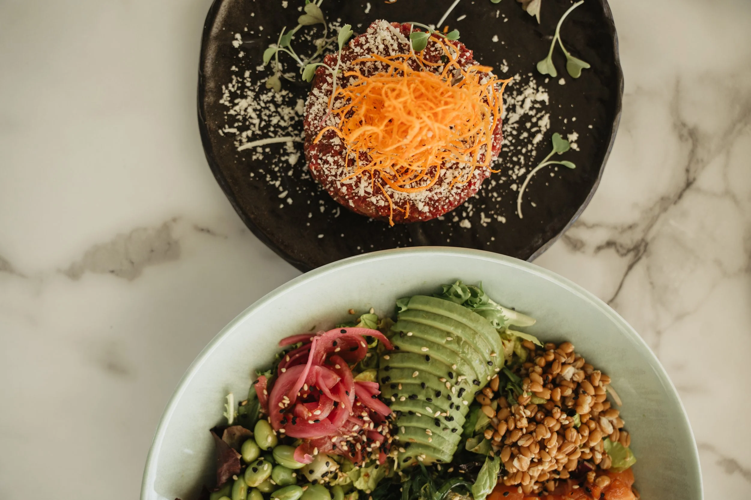 A salad with sliced avocado, pickled onions, beans, and mixed greens in a white bowl. A separate dish with a topped veggie burger or meatball, shredded carrots, microgreens, and grated cheese on a black plate.