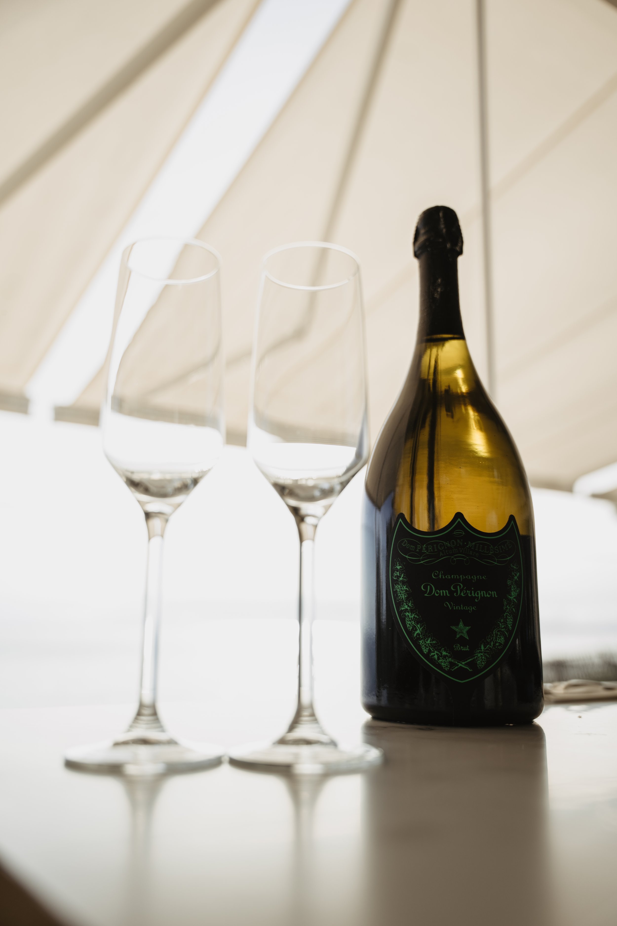 A bottle of Dom Pérignon champagne and two empty champagne flutes on a white surface with a tent-like structure overhead.