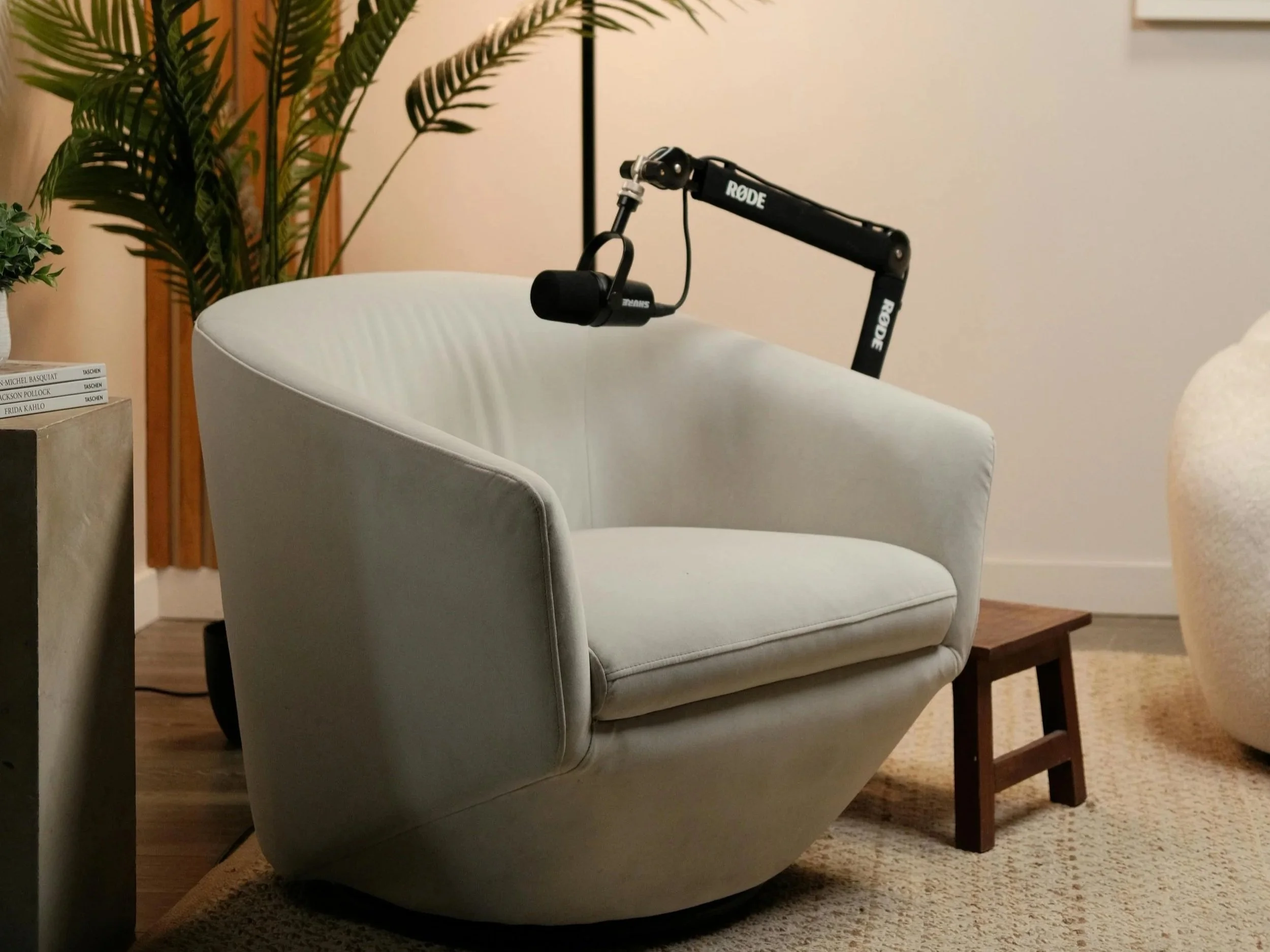 A white armchair with a mounted black Rode microphone arm attached to its armrest, situated next to a small wooden stool and a concrete side table with books. There are green plants and a tall lamp in the background, in a cozy living room setting.