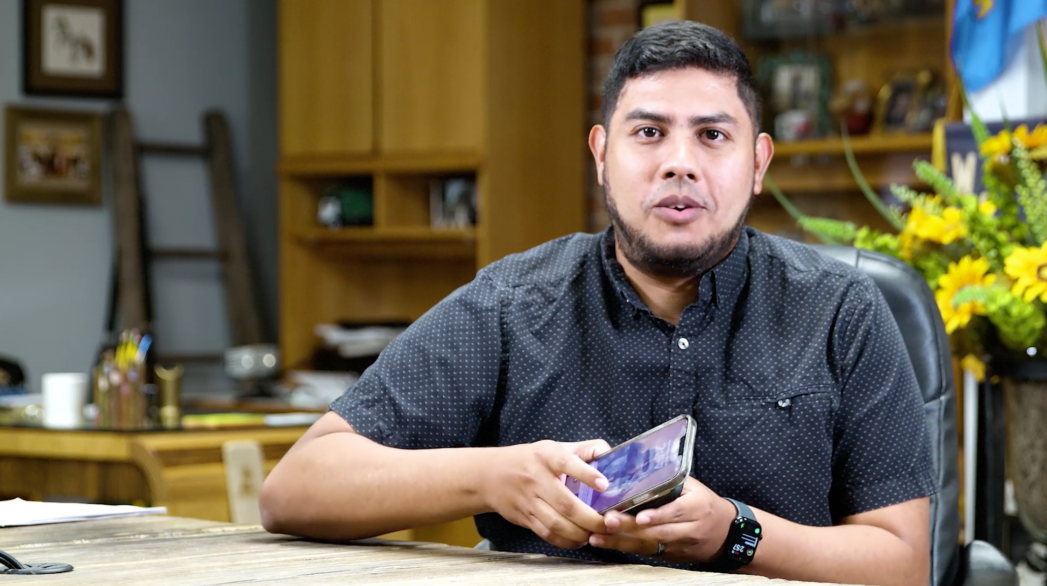 A man sitting at a desk holding a smartphone, with a background of bookshelves and a vase of yellow flowers.