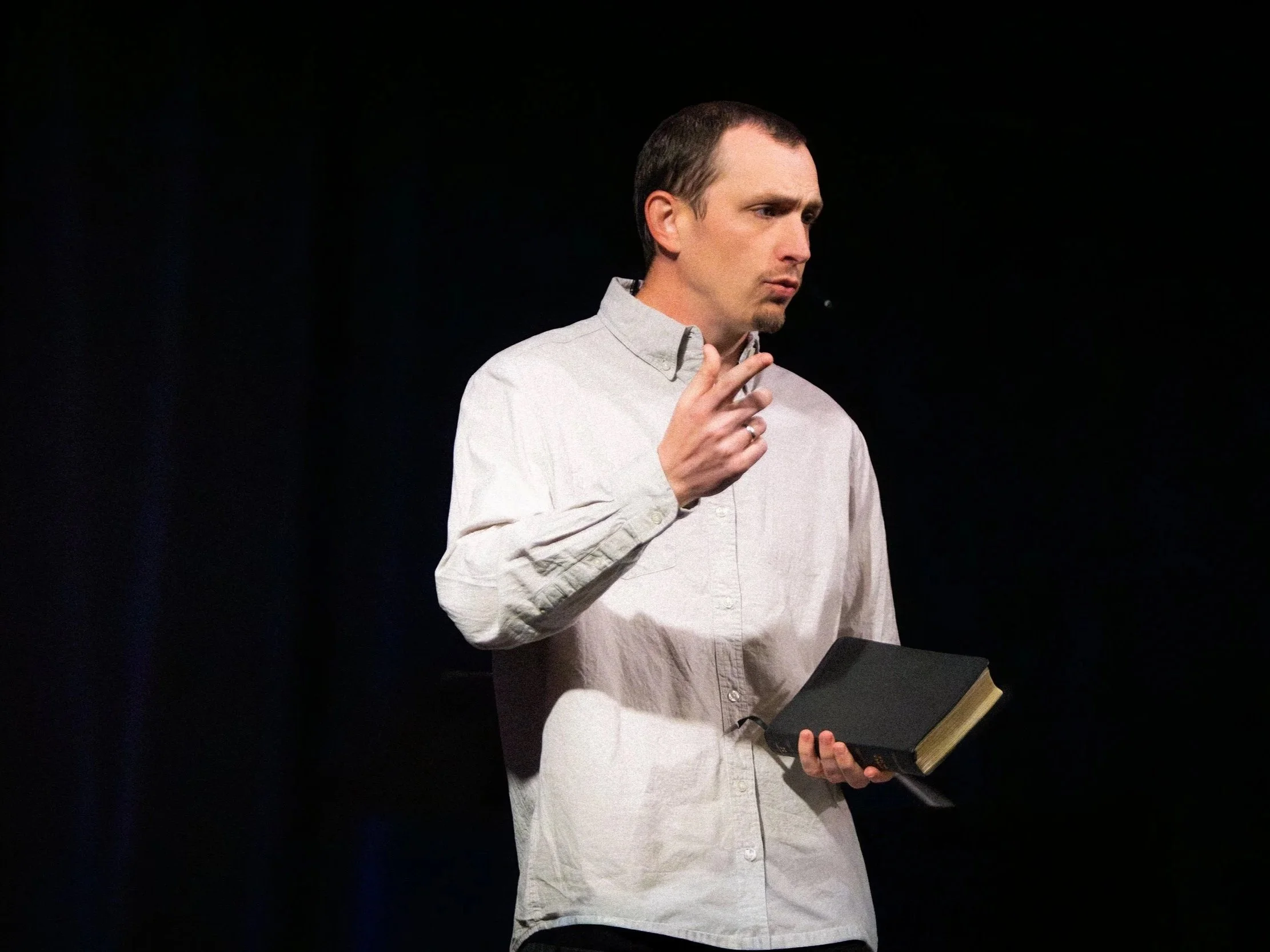 A man with short dark hair, wearing a light gray button-up shirt, is holding a black book and making a directing gesture with his right hand, against a plain dark background.