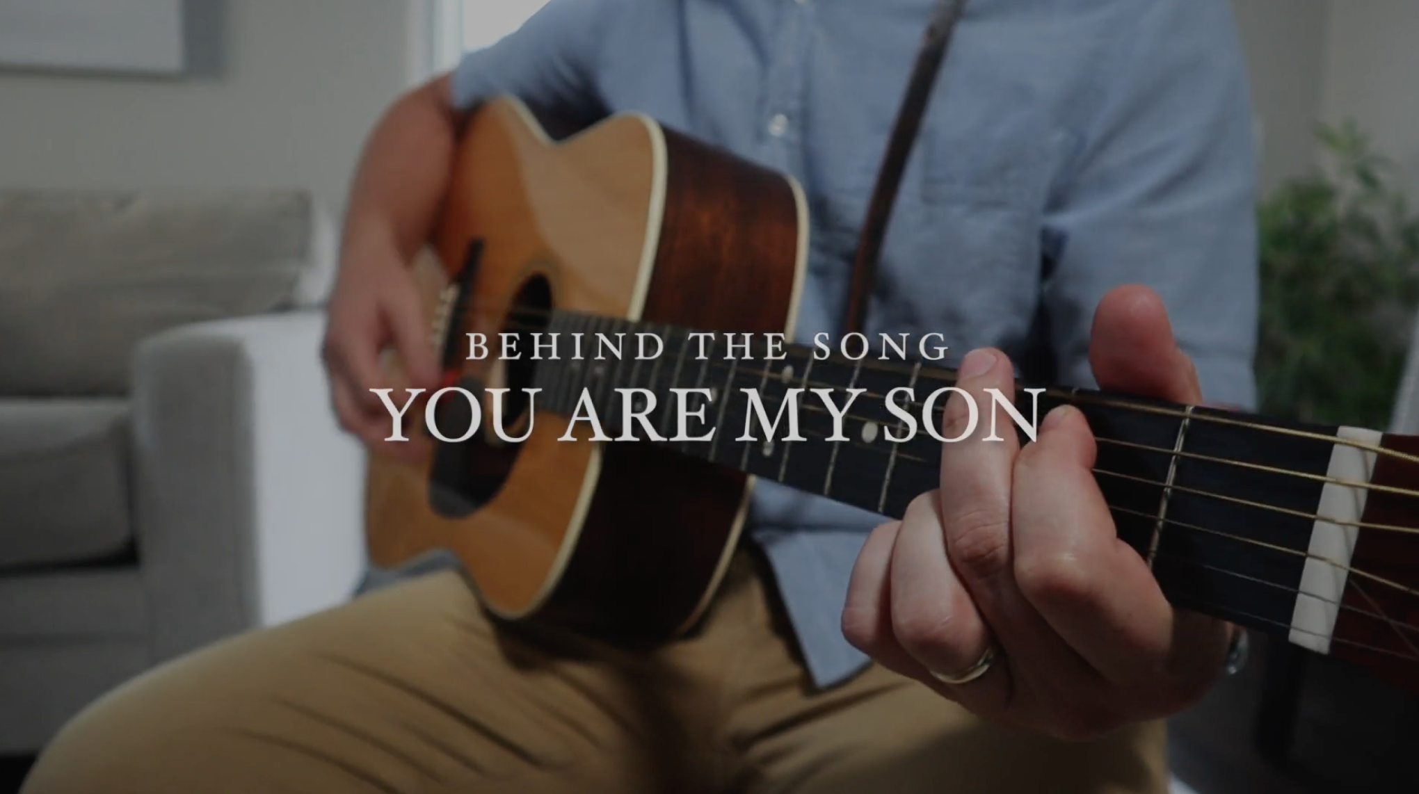 A person playing an acoustic guitar with text overlay reading 'Behind the song You Are My Son' in a cozy living room setting.