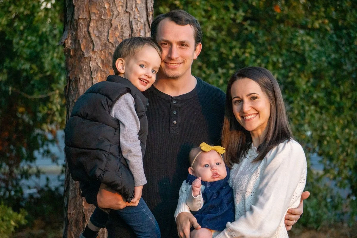 Family portrait of a father, mother, and two young children smiling outdoors next to a tree, with green foliage in the background.