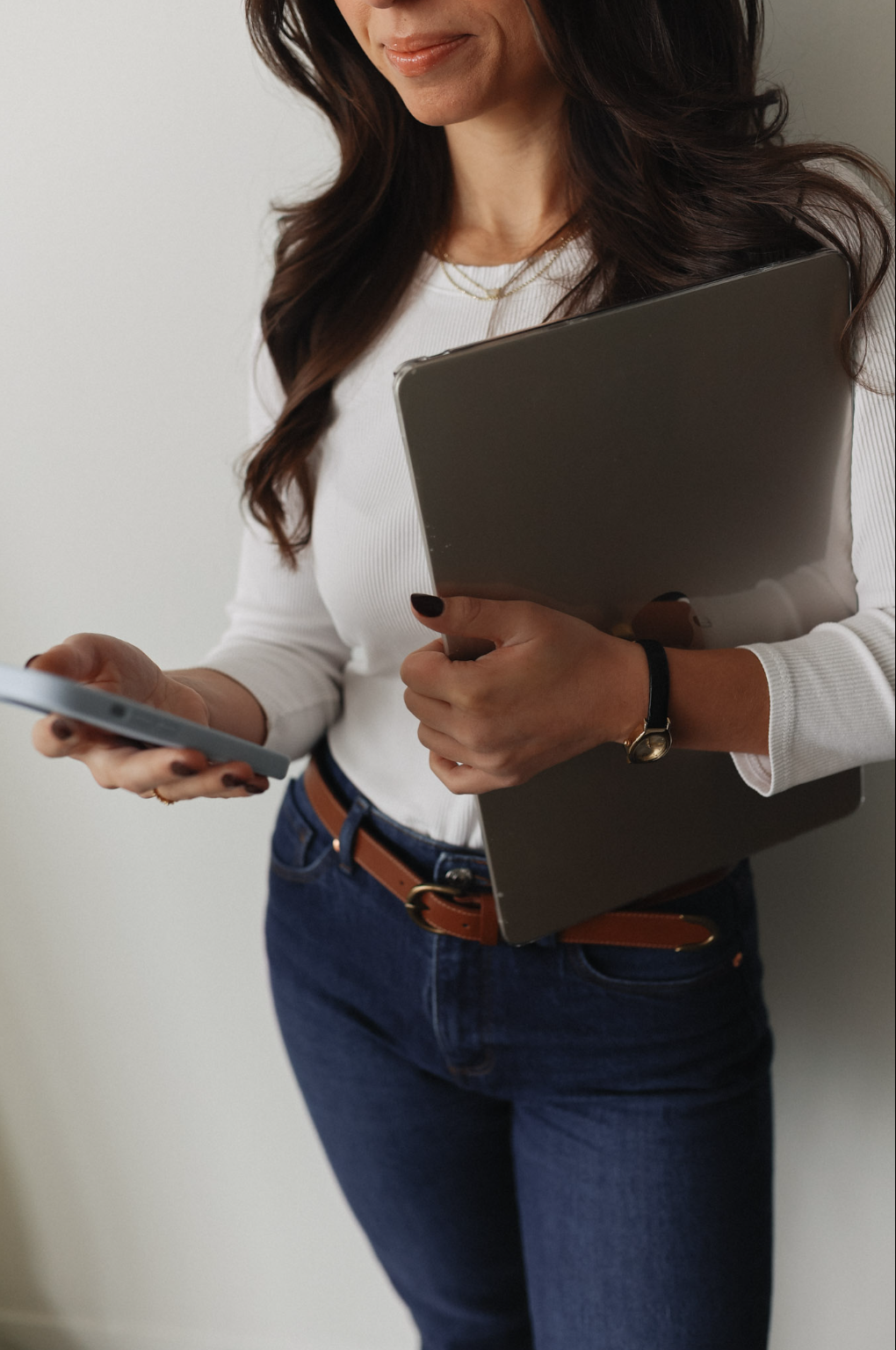 A woman holding a laptop and a smartphone, dressed in a white top and jeans, standing against a plain wall.