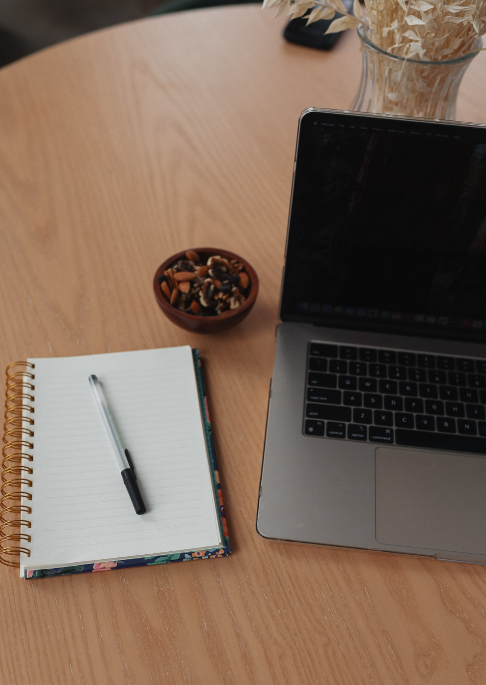 A workspace on a wooden table with a laptop, an open blank spiral notebook with a pen, a bowl of trail mix, and a vase of dried flowers.