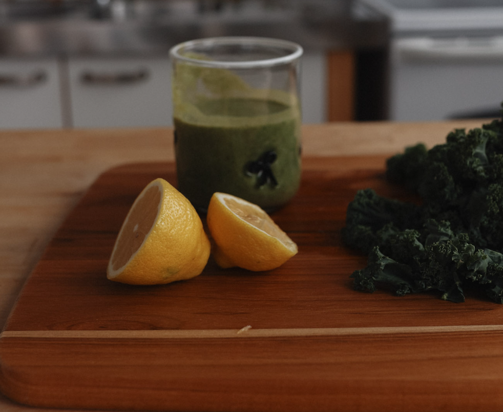 Lemon halves, a glass of green smoothie, and kale on a wooden cutting board in a kitchen.