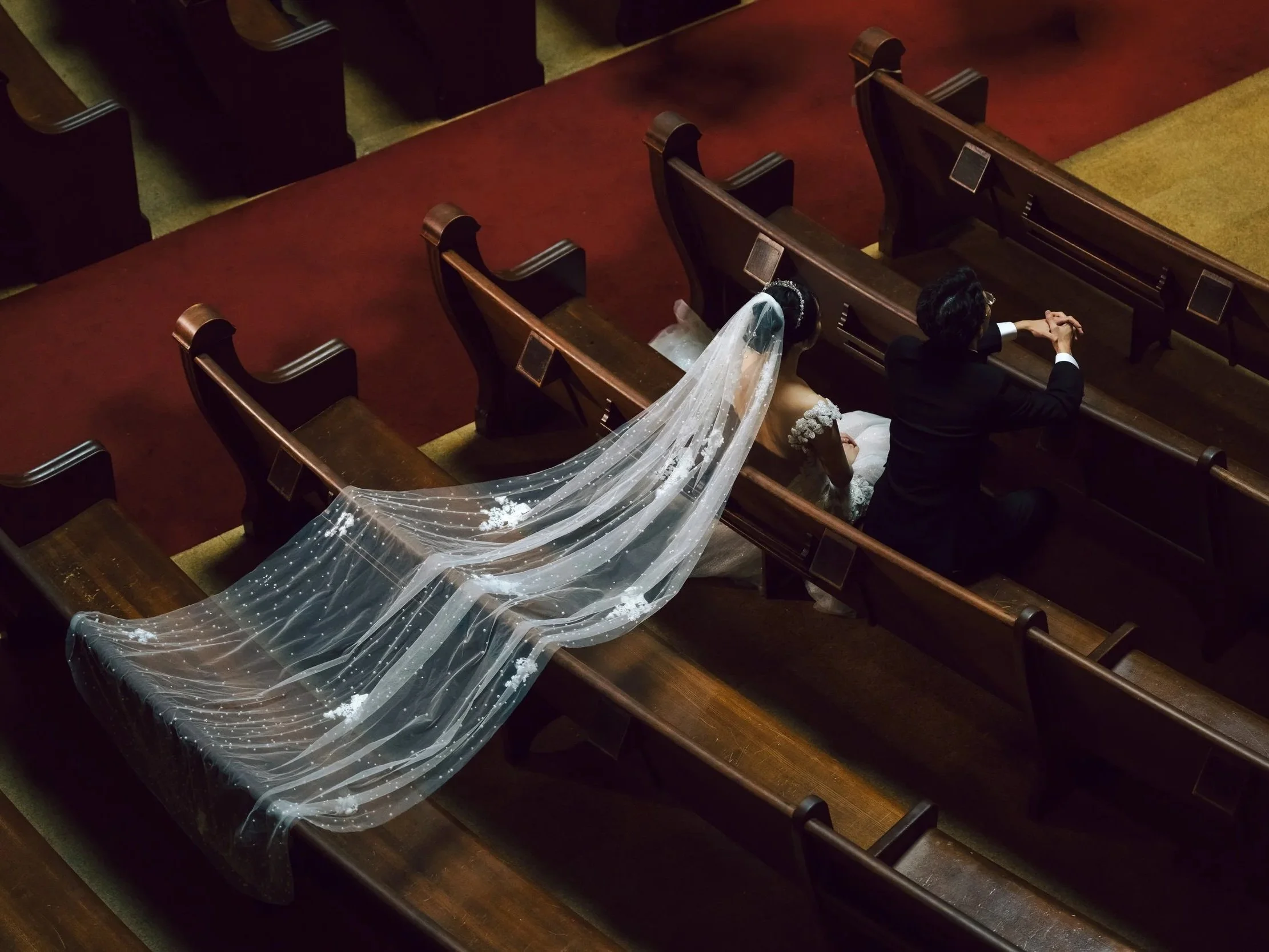 Top view of a bride and groom sitting in church pews, holding hands, with the bride wearing a wedding gown and veil, and the groom in a black suit. The bride's long veil extends across empty pews.