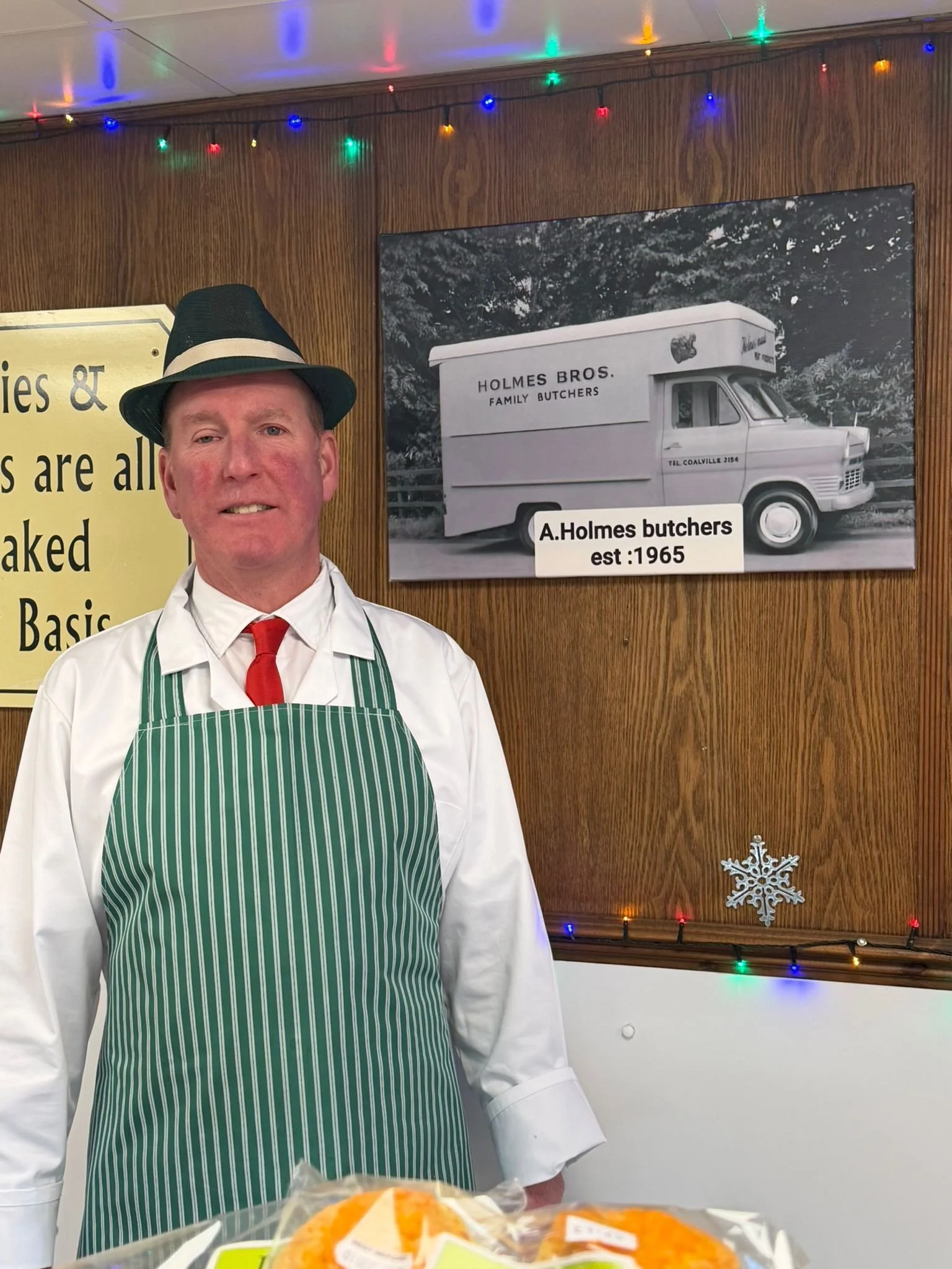 A man wearing a black fedora, white shirt, red tie, and green striped apron stands indoors with wooden paneling and colorful holiday lights. Behind him is a black-and-white photo of a vintage food truck with sign "Holmes Bros. Family Butchers est: 1965". Part of a yellow sign and a snowflake decoration are also visible.