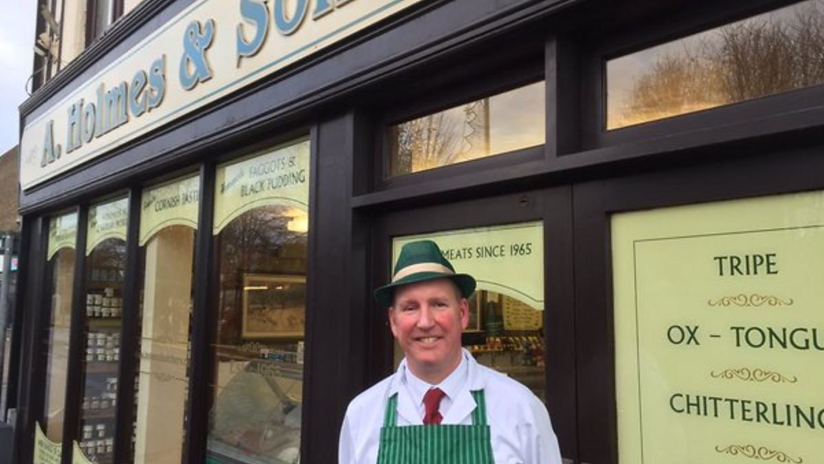 A man wearing a green hat and apron, standing outside a butcher shop with signs advertising various meats.