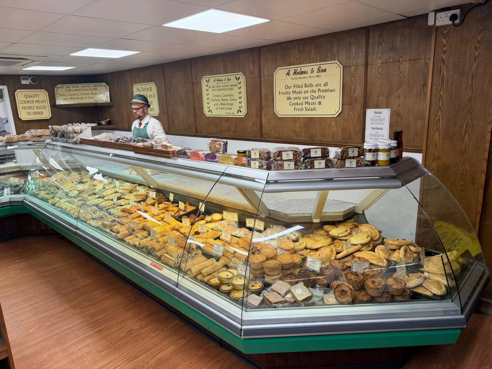 A bakery display case filled with various baked goods such as pastries, tarts, and cookies, with a man in a striped apron and hat standing behind the counter. There are signs on the wooden wall behind him advertising quality meats and other products.