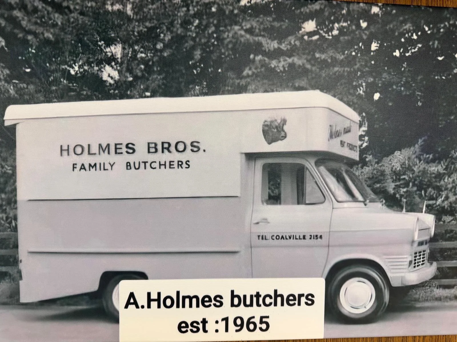 A vintage Holmes Bros. family butcher truck parked outdoors, with trees in the background. The truck has the company name and a logo on the side, along with the phone number 'TIL COALVILLE 2148'. There is a label at the bottom that reads 'A. Holmes butchers est: 1965'.