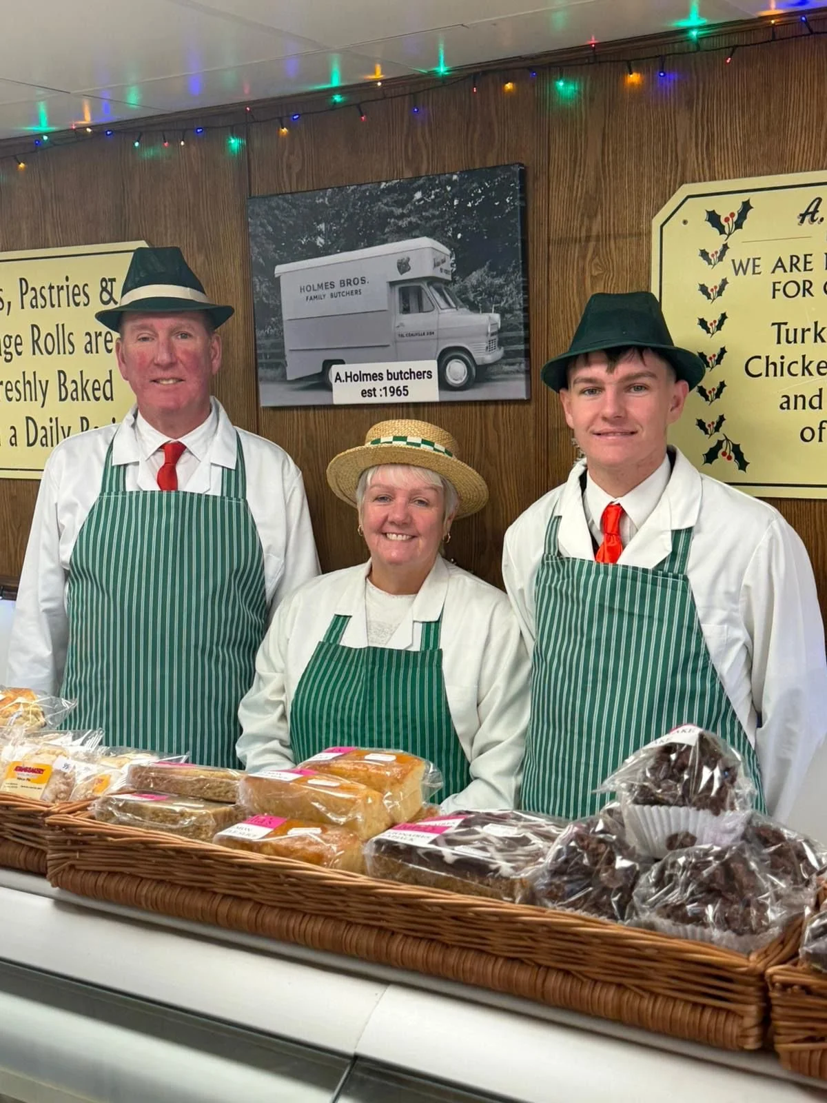 Three people in vintage costume standing behind a display of baked goods at a bakery counter, with signs and photos on the wall behind them.