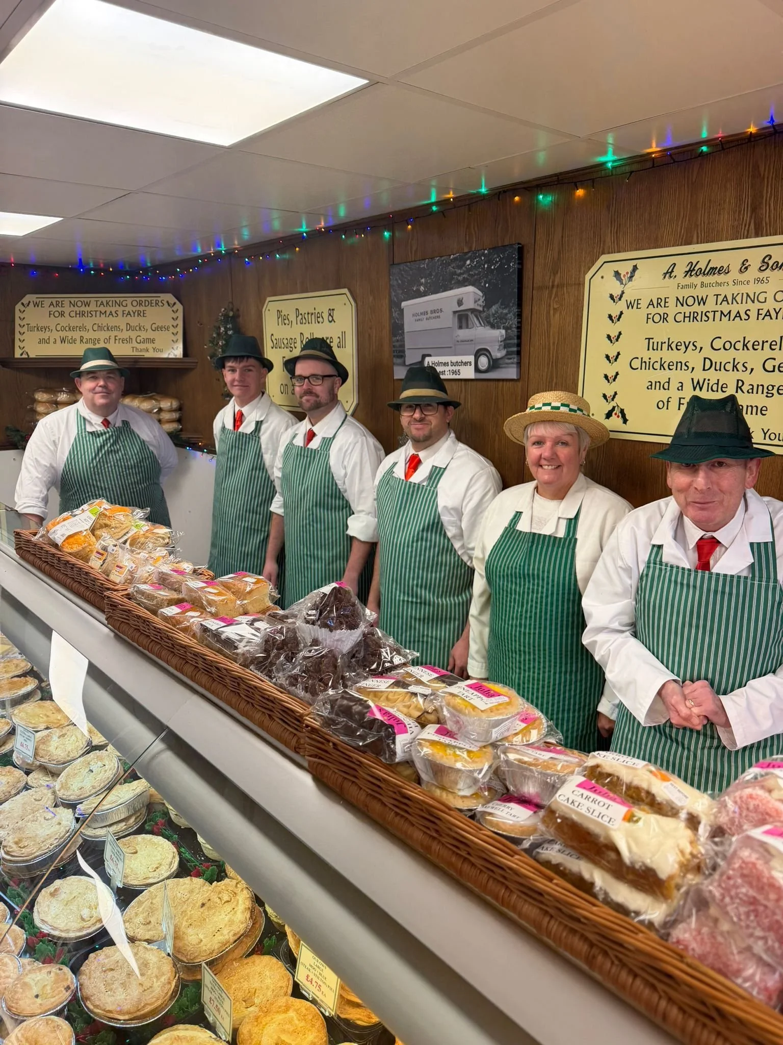 Six people dressed in white shirts, green aprons, and hats standing behind a bakery counter with baked goods. The bakery has signs and Christmas lights on the walls.