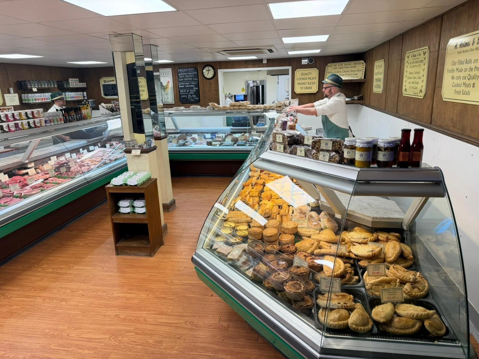 Inside a butcher shop, a man wearing a white shirt, apron, and glasses arranges meat and baked goods on display counters. The shop has wooden floors and walls with signage about products and services. There are glass display cases with various meats, baked goods, and condiments, as well as shelves with additional items.