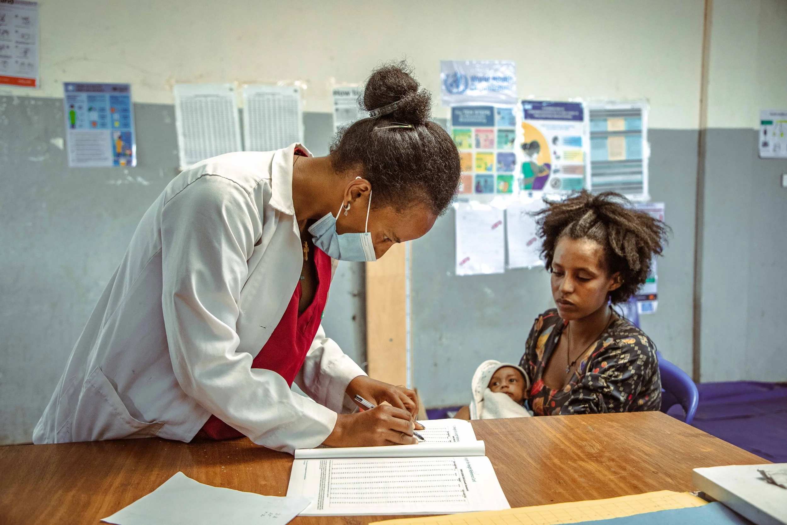 A healthcare worker in a surgical mask and white coat is signing a document on a wooden table, with a woman holding a baby seated nearby in a makeshift clinic or registration area with informational posters on the wall.