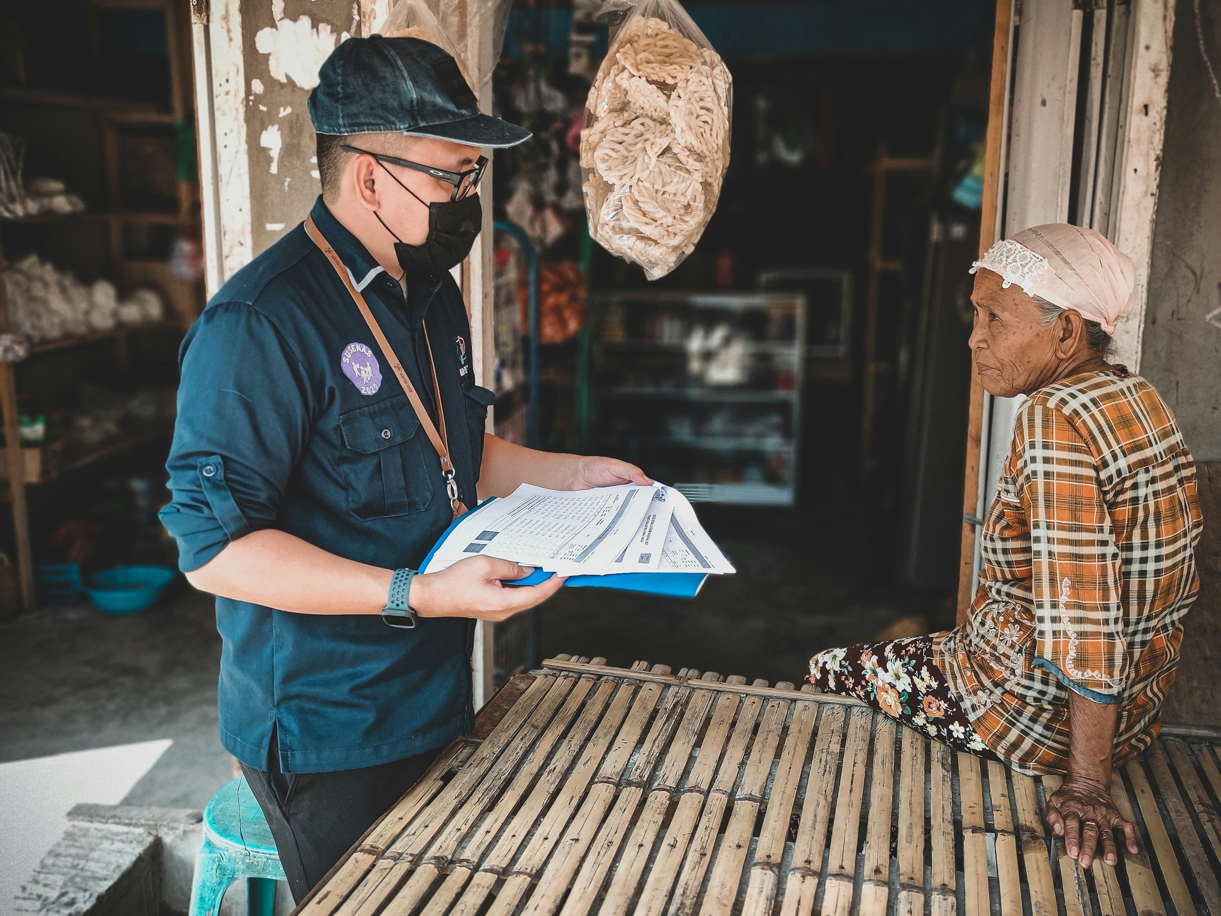 A man wearing a black mask, glasses, and a dark blue shirt with patches is holding papers and talking to an elderly woman sitting on a wooden table outside a small shop.