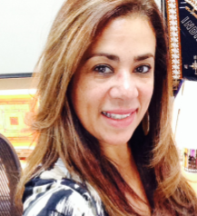 A smiling woman with dark curly hair wearing a purple sweater stands in front of green leafy bushes.