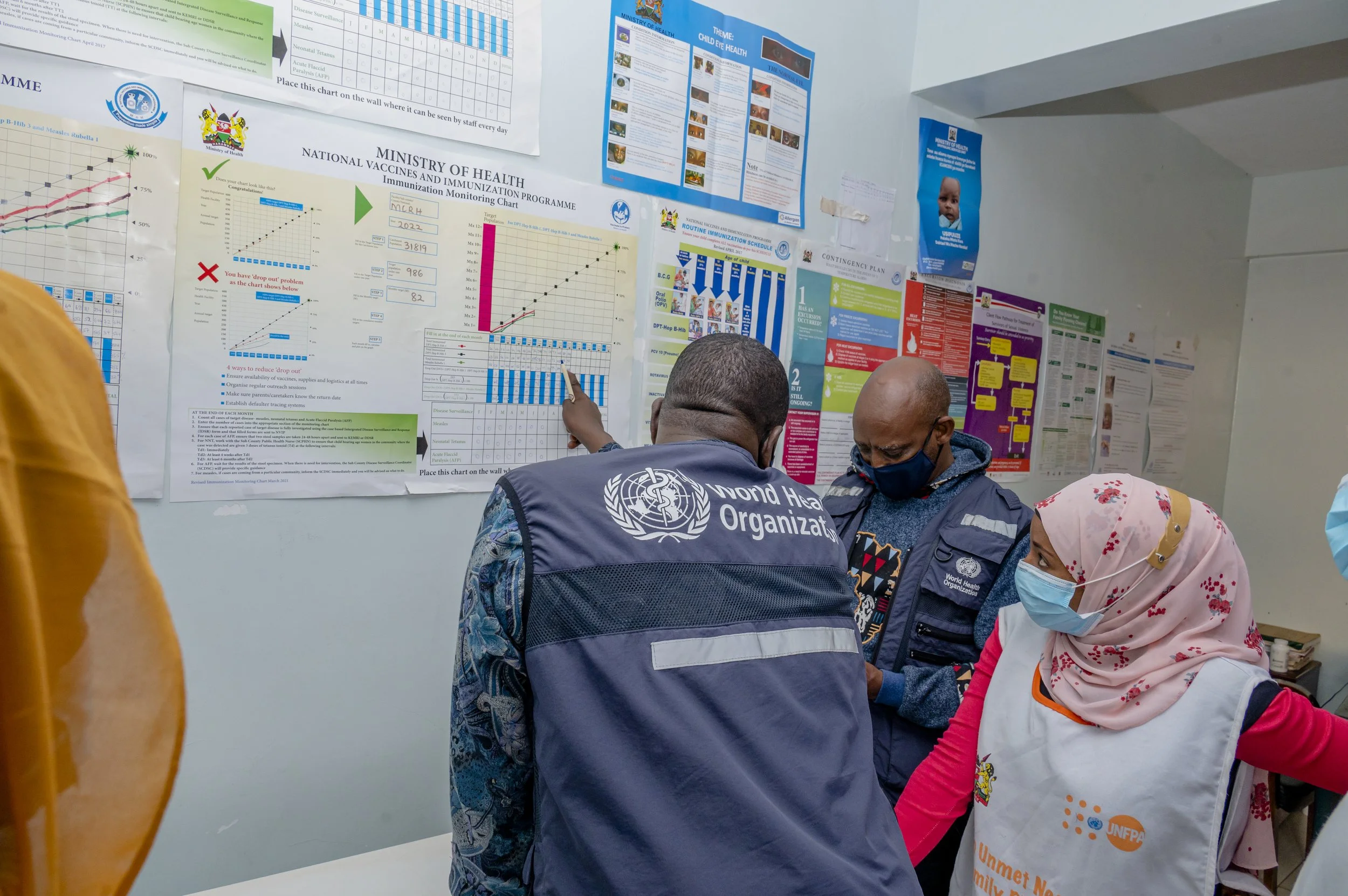 Health officials from WHO and UNICEF discuss vaccination data and schedules on a wall chart at an indoor health setting, with informational posters on child health and immunization visible in the background.
