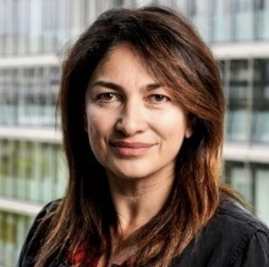 A woman with shoulder-length brown hair, wearing a black blazer, standing outdoors in front of a modern office building with glass windows.