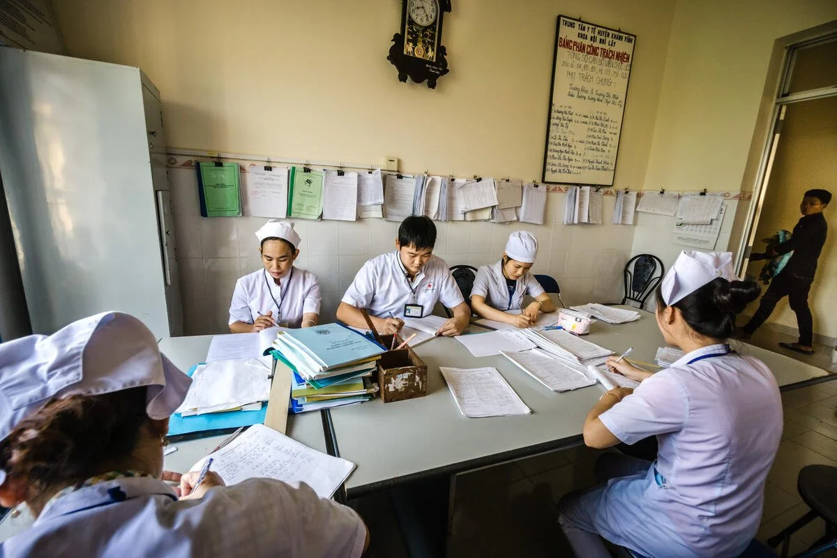 Group of healthcare professionals, including nurses in uniform and caps, sitting at a table with files, papers, and writing supplies, in a medical or hospital office.