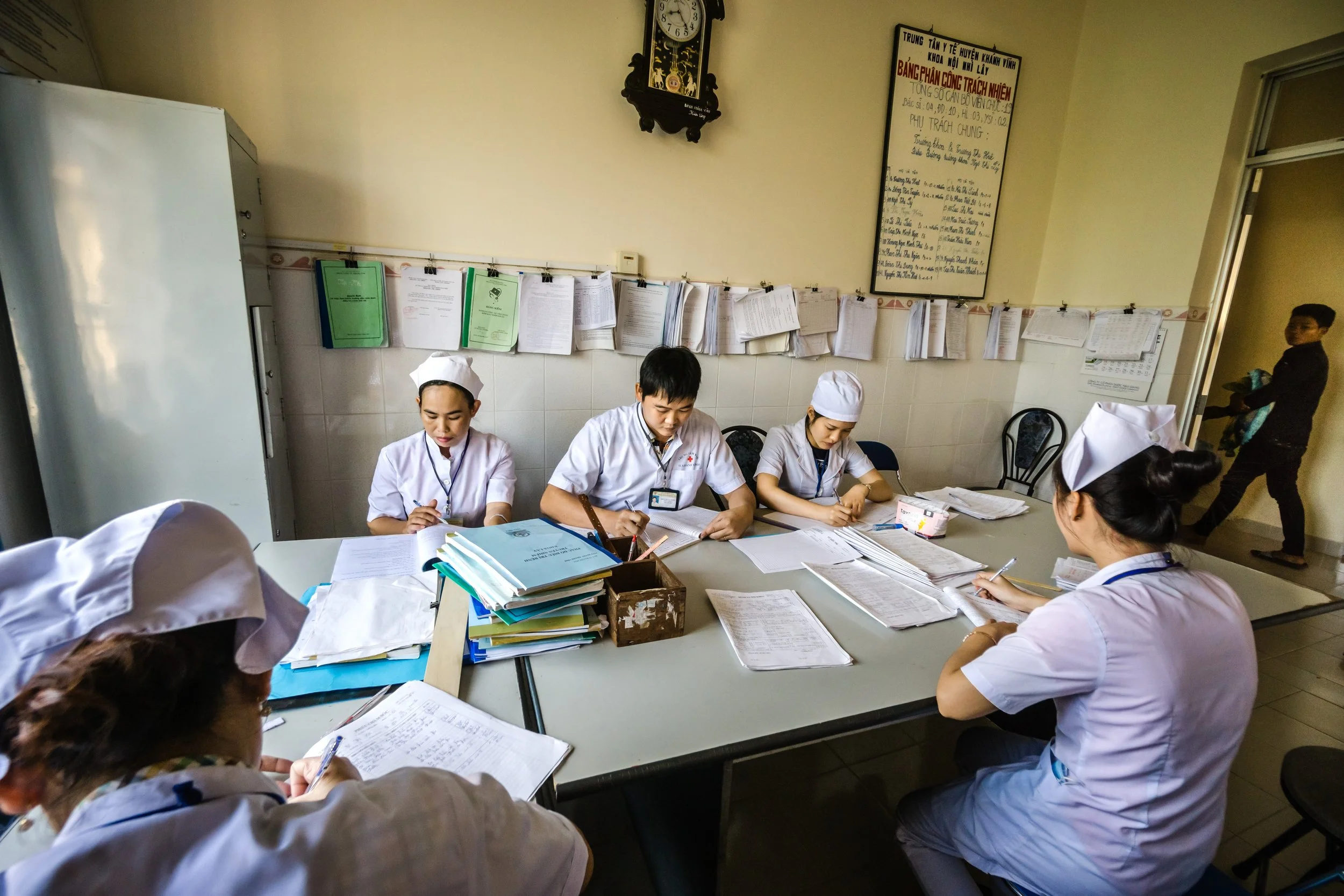 Healthcare workers wearing white uniforms and nurse caps sitting around a table filled with paperwork, studying or discussing medical documents in a room with paperwork on the wall and a clock.
