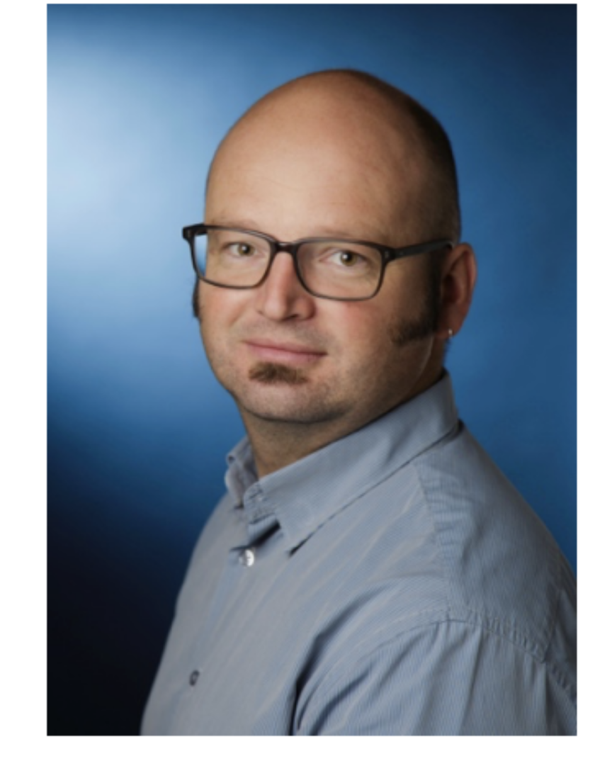A portrait of a middle-aged man with glasses and a goatee, wearing a light blue collared shirt, against a blue gradient background.