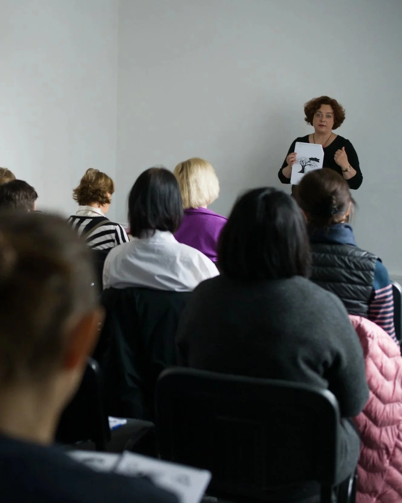 A woman giving a presentation to a group of people in a conference room. The presenter is holding a paper with a tree illustration.