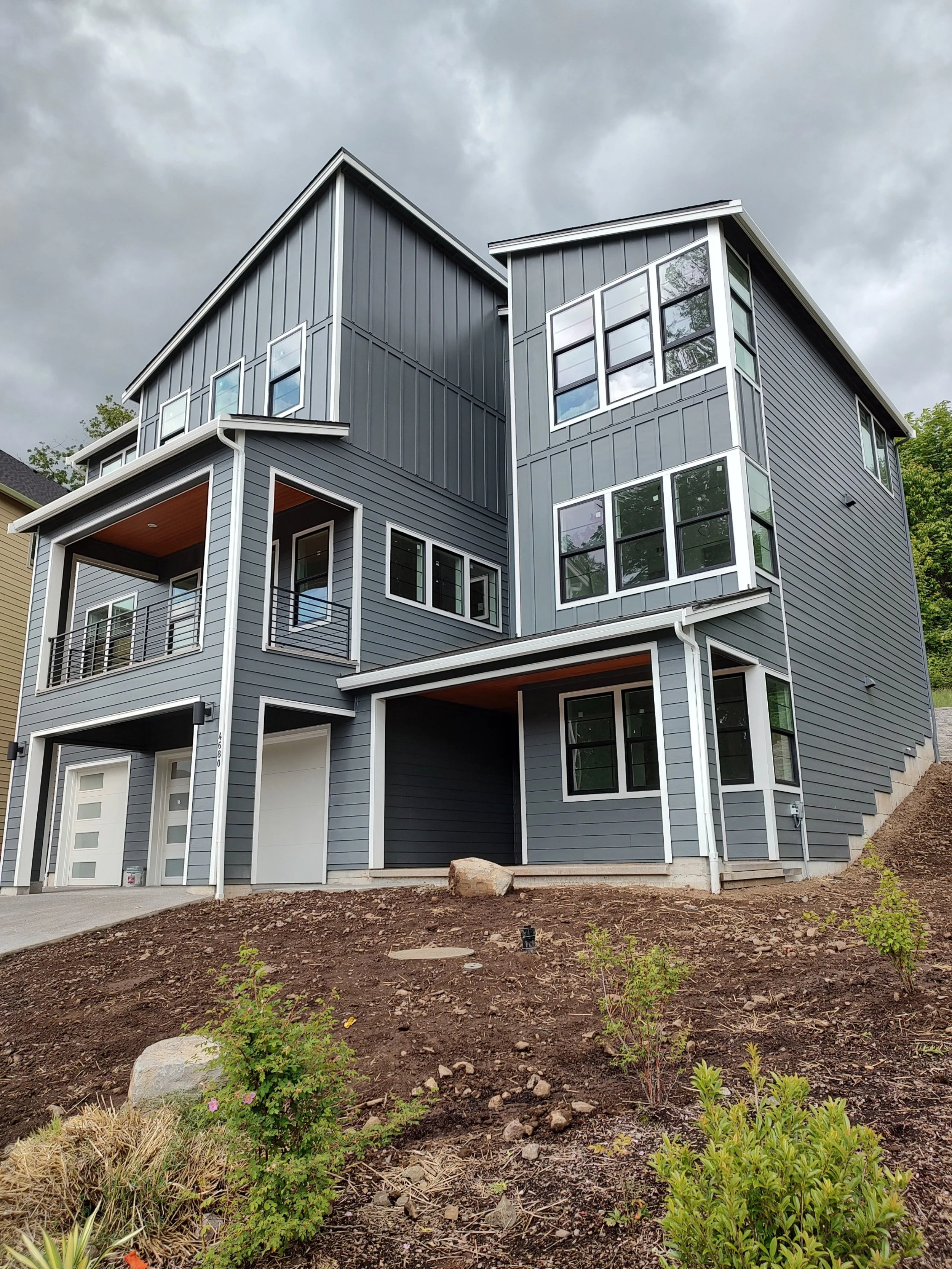 New modern multi-story house with gray exterior, large windows, and a small balcony, situated on a sloped lot with landscaping and a cloudy sky
