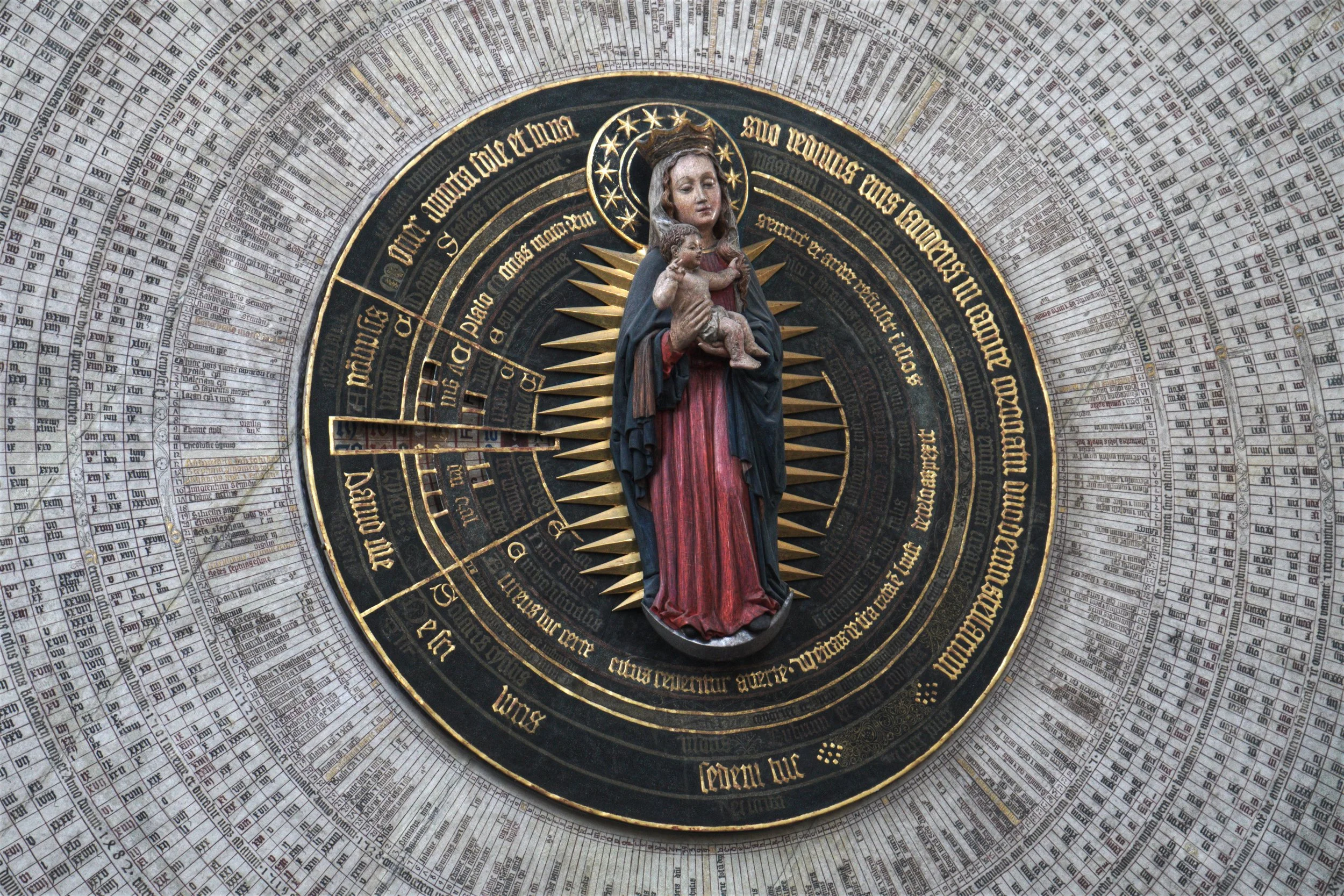 Religious clock with Madonna and Child sculpture at the center surrounded by Latin text and time markers.