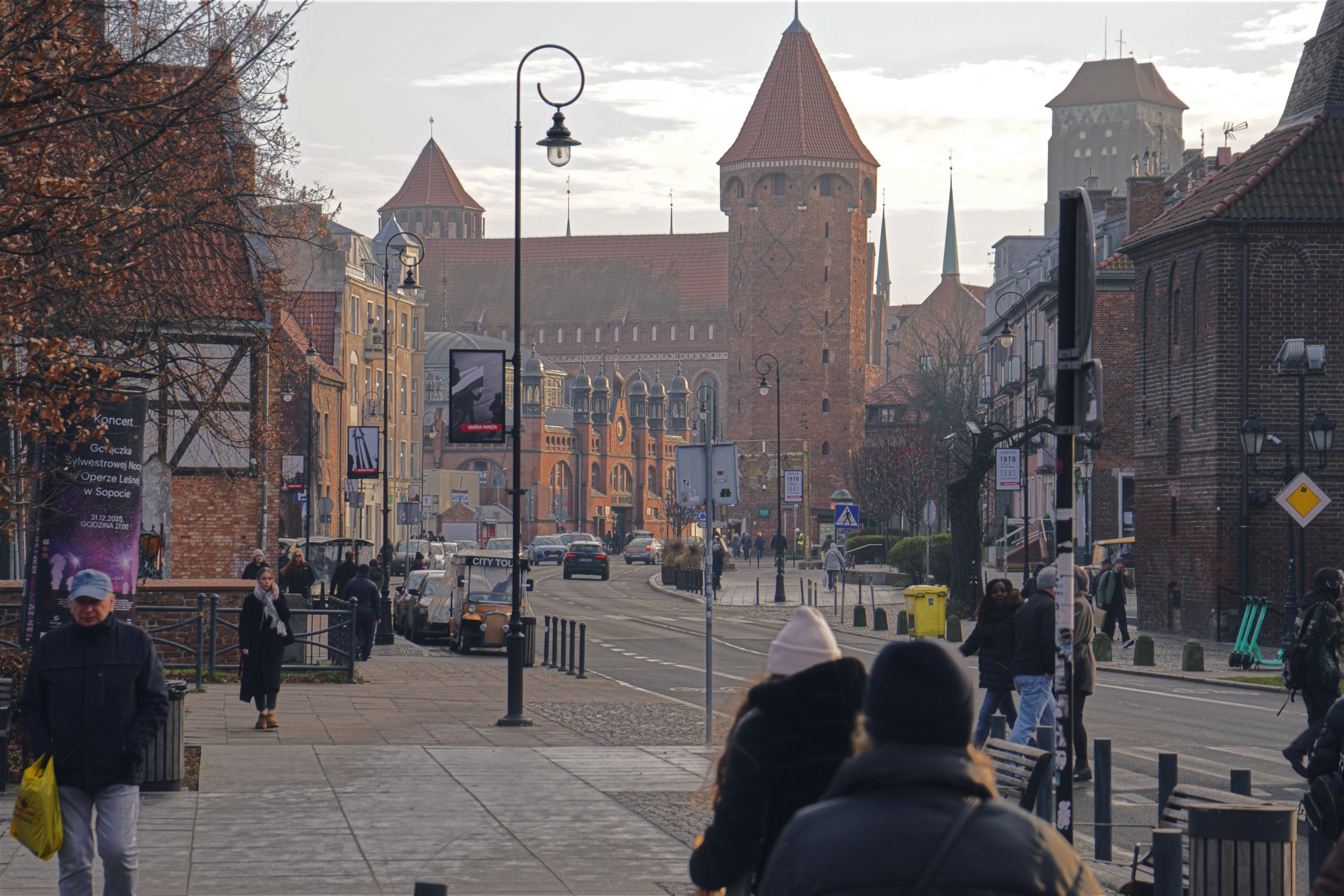 Street scene in a historic European city, with pedestrians walking along a sidewalk, old brick buildings, street lamps, and a large medieval brick church with towers in the background.