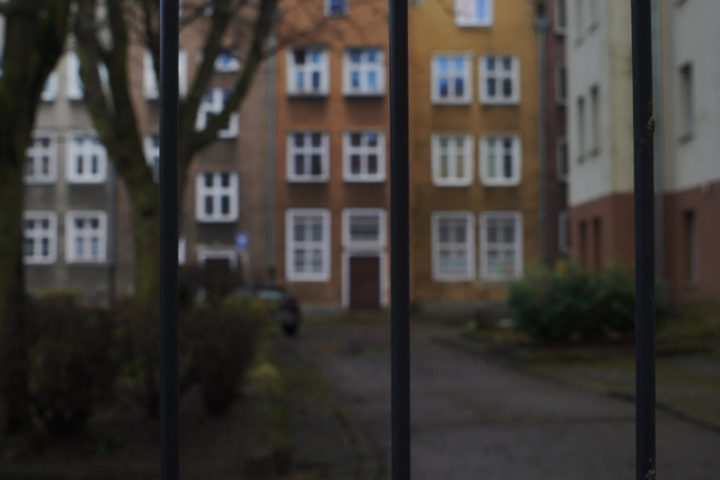 View of residential apartment buildings seen through black metal bars, with trees and shrubbery in the foreground, during daytime.