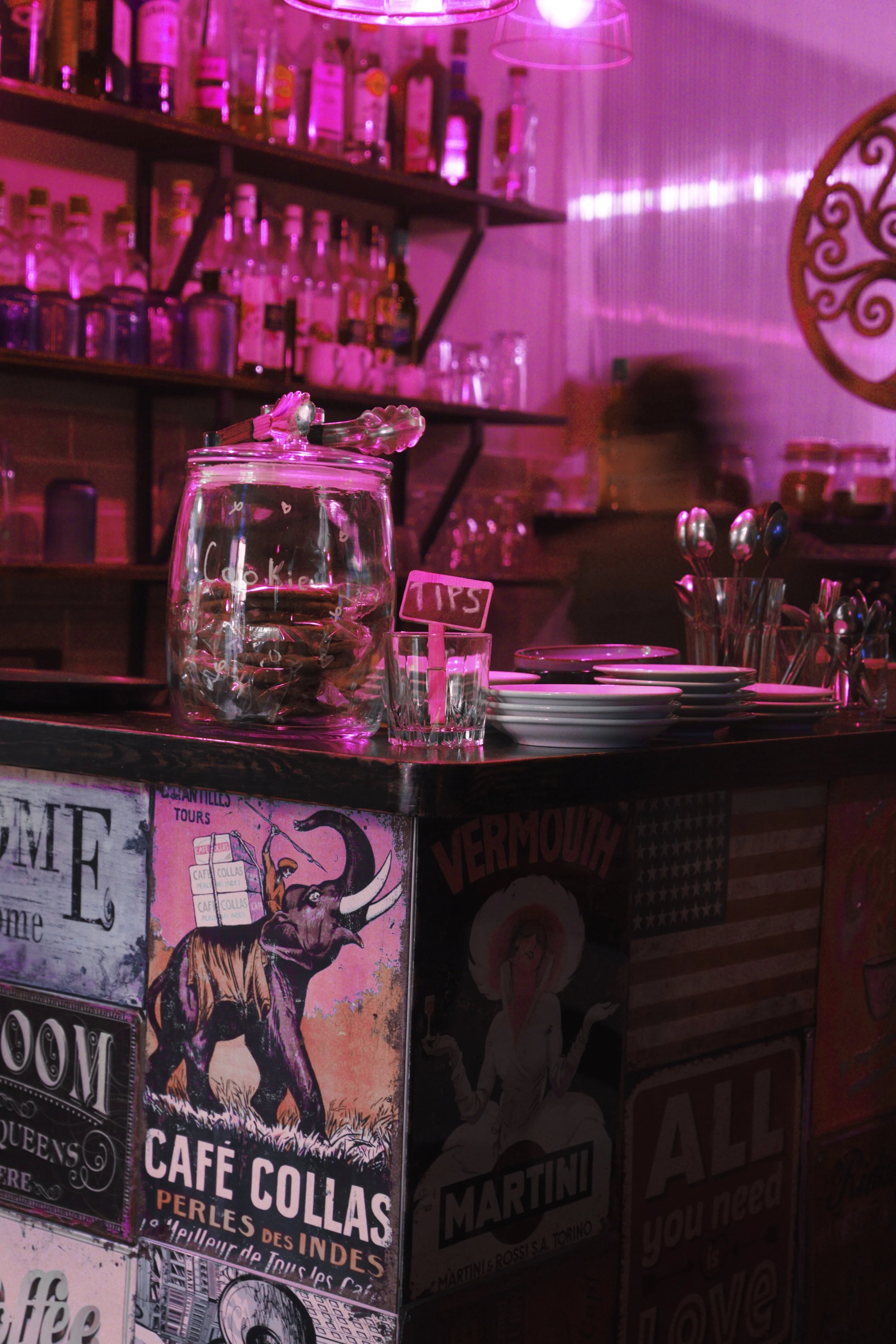 Bar counter with glass jar labeled 'Cookies', a small tip jar, stacked plates, and utensils, with shelves of bottles and glasses behind, and vintage style posters on the front.
