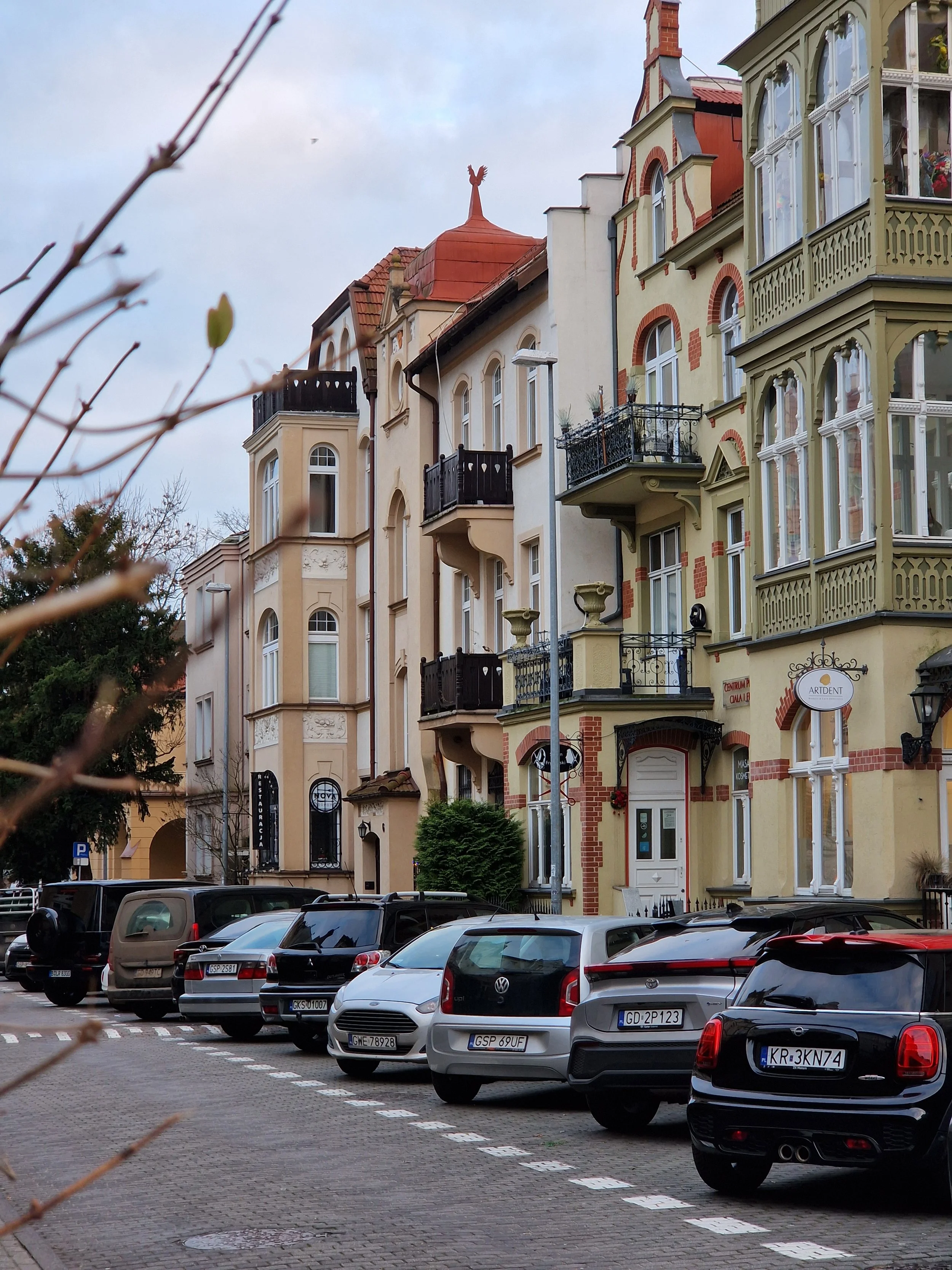 Colorful old European-style apartment buildings with bay windows, balconies, and decorative architectural details, parked cars in the foreground, overcast sky.