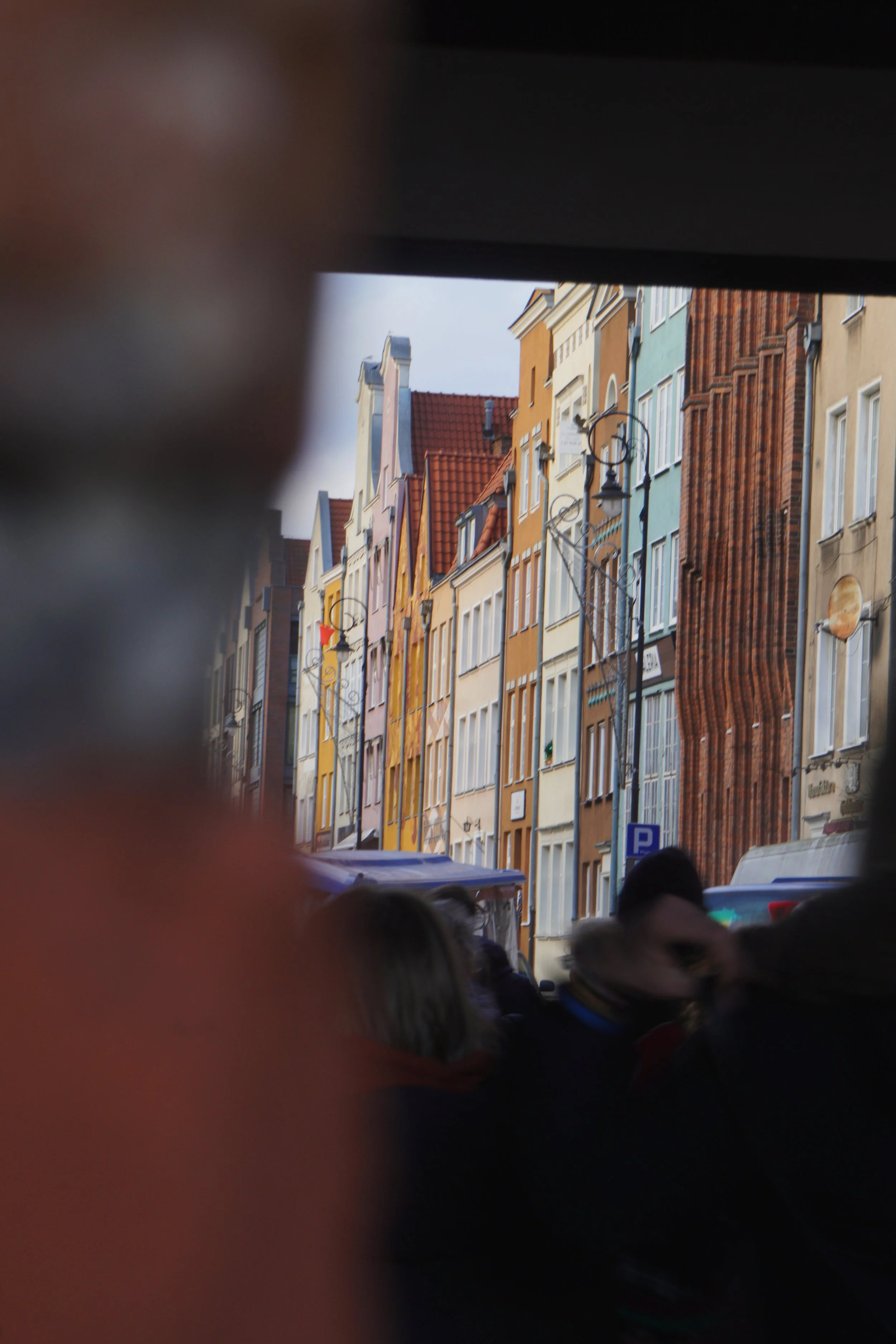 Colorful row of multi-story buildings with traditional European architecture, seen through a small opening among people in the foreground.