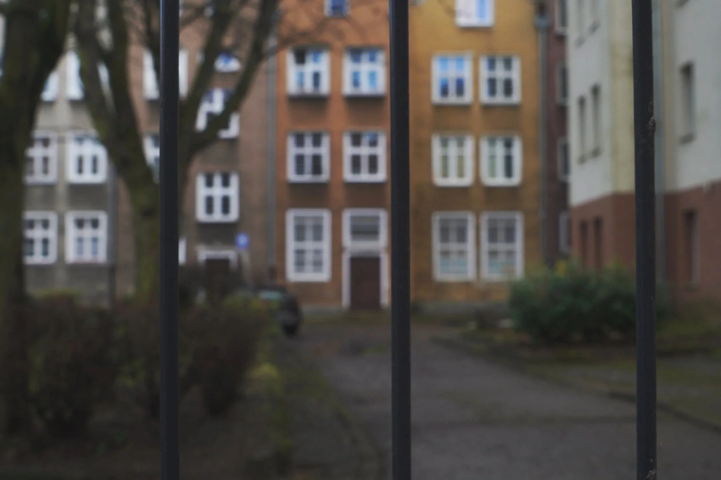 Blurred view of an apartment complex seen through metal bars, with trees and shrubs in the foreground.