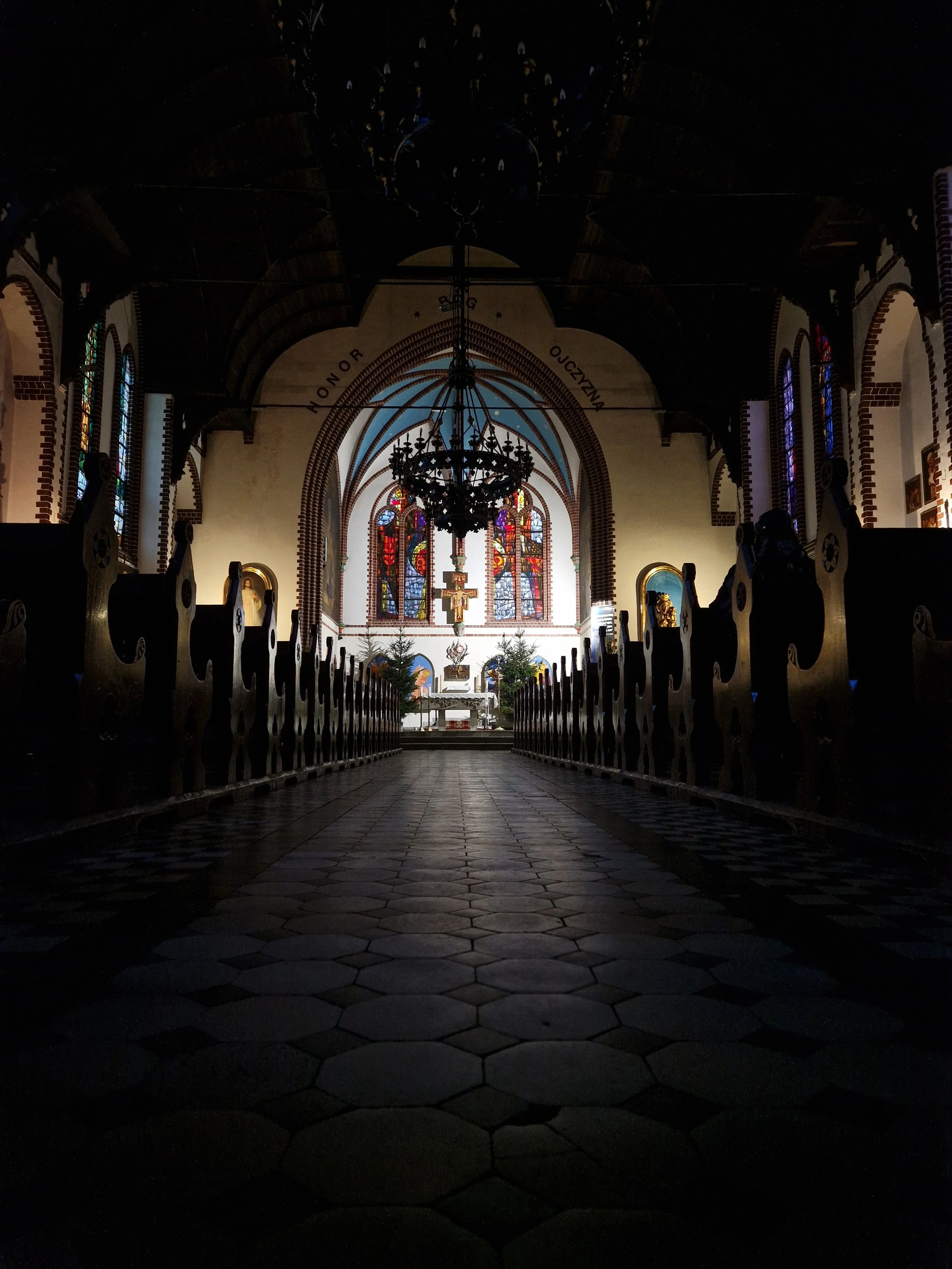 Interior view of a church with a central aisle, dark pews, stained glass windows, a large chandelier, and an altar at the front with religious icons and Christmas trees.
