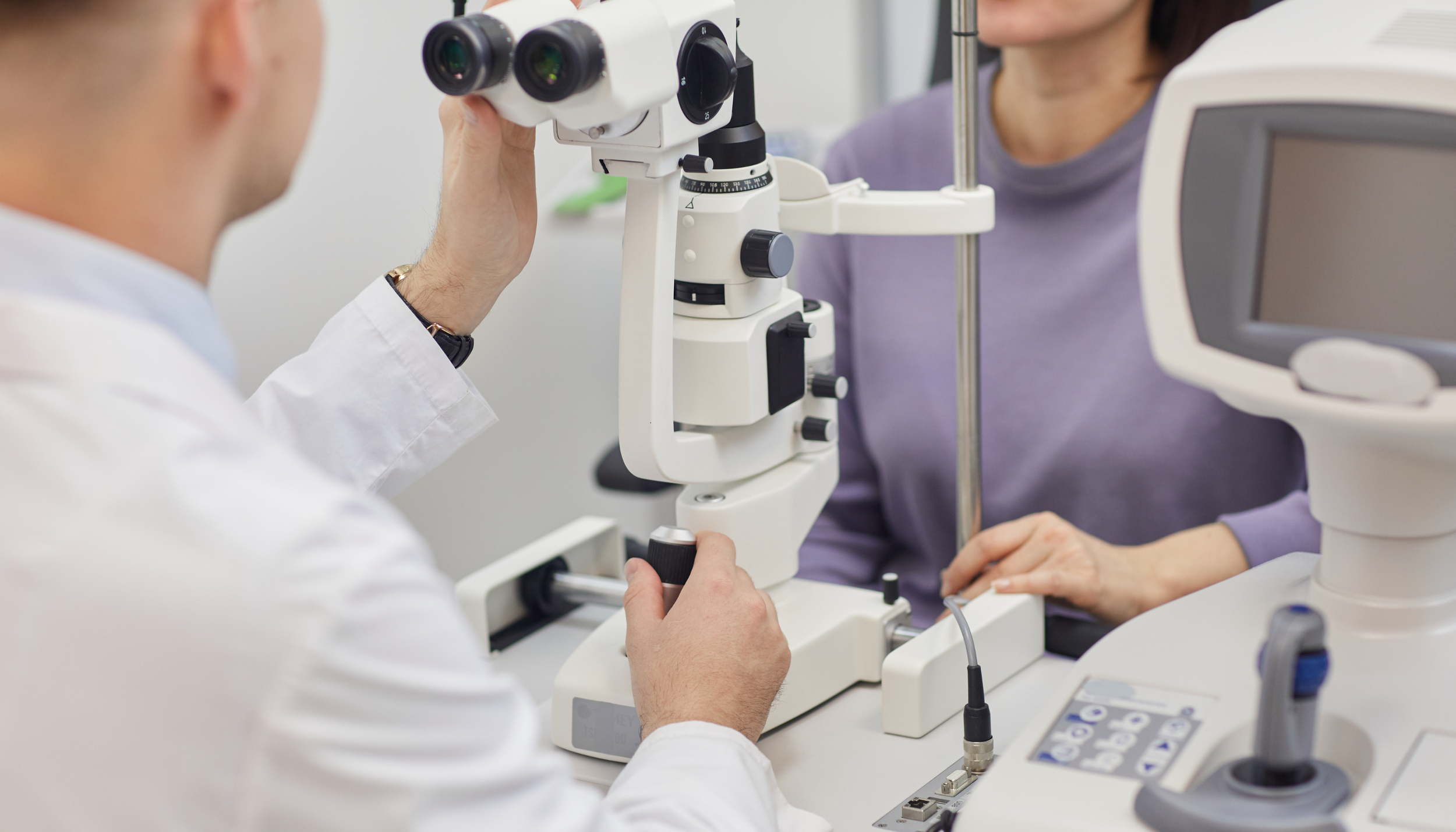 A healthcare professional in a white coat using a microscope during an eye examination of a patient seated at the desk. The patient is wearing a purple shirt and is looking through the microscope.