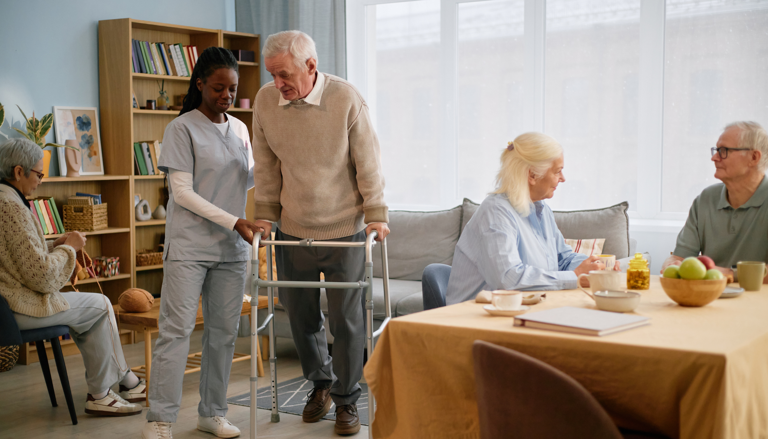 An elderly man with white hair using a walker in a room surrounded by four women, one of whom is a healthcare worker assisting him. The women are sitting and standing around a wooden table with breakfast items, in a bright room with large windows and a bookshelf.
