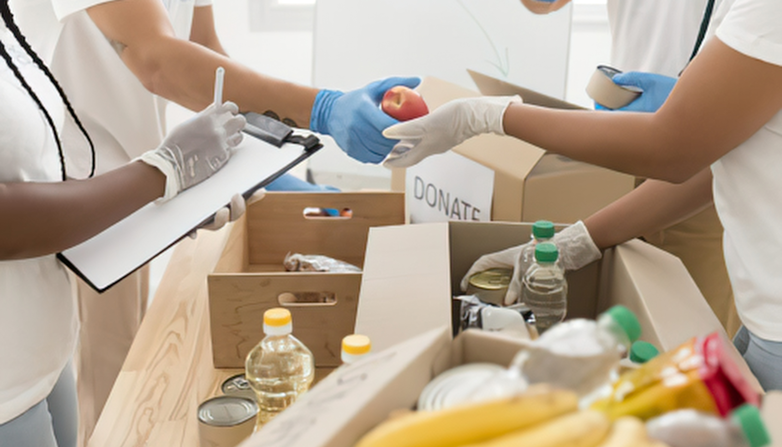 People in white shirts donating food and drinks, with a donations box and bottles of water in a bright room.